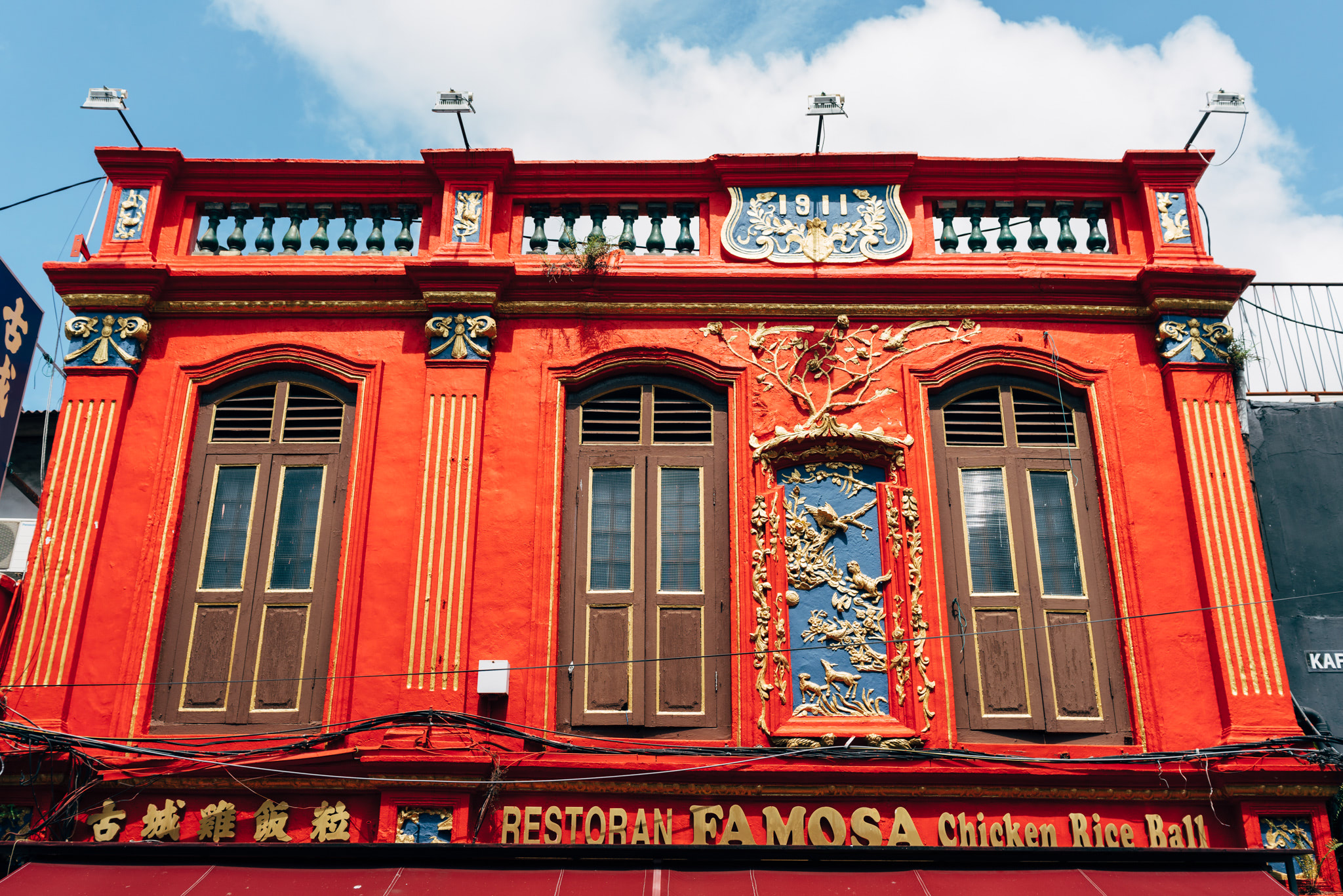 Bright red building with ornate gold details, three dark brown windows, and a sign that says 'Restoran Famosa Chicken Rice Ball'.