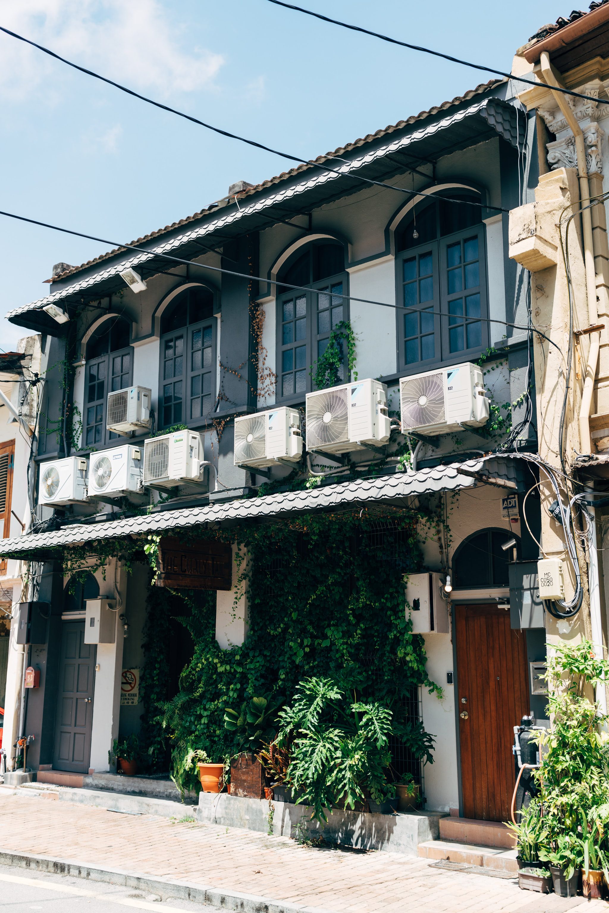 A two-story building in Malacca, Malaysia, with multiple air conditioning units mounted on the exterior wall, and lush green plants growing at the base.