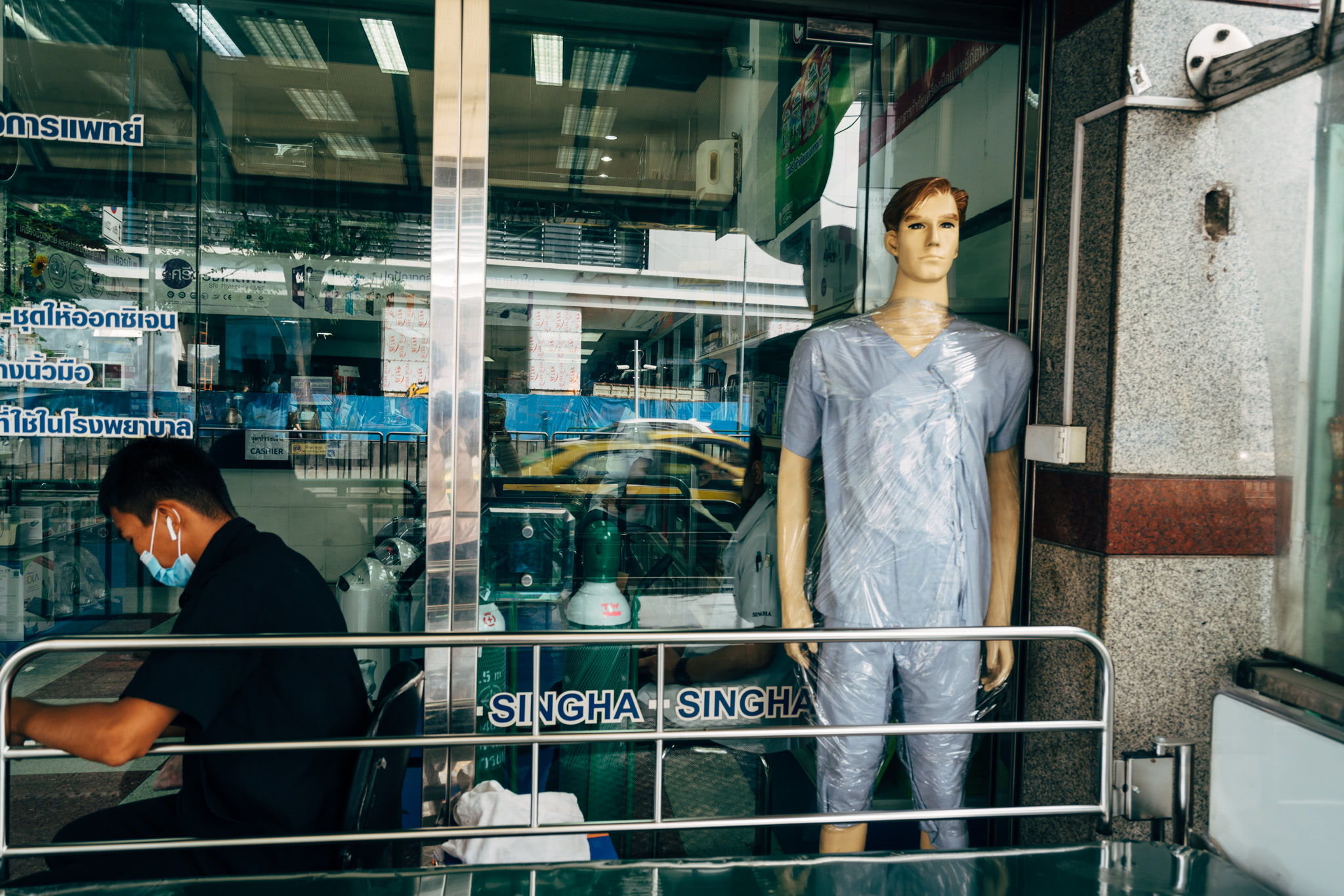Bangkok street scene: a mannequin in scrubs stands by a storefront, a person wearing a mask sits nearby.