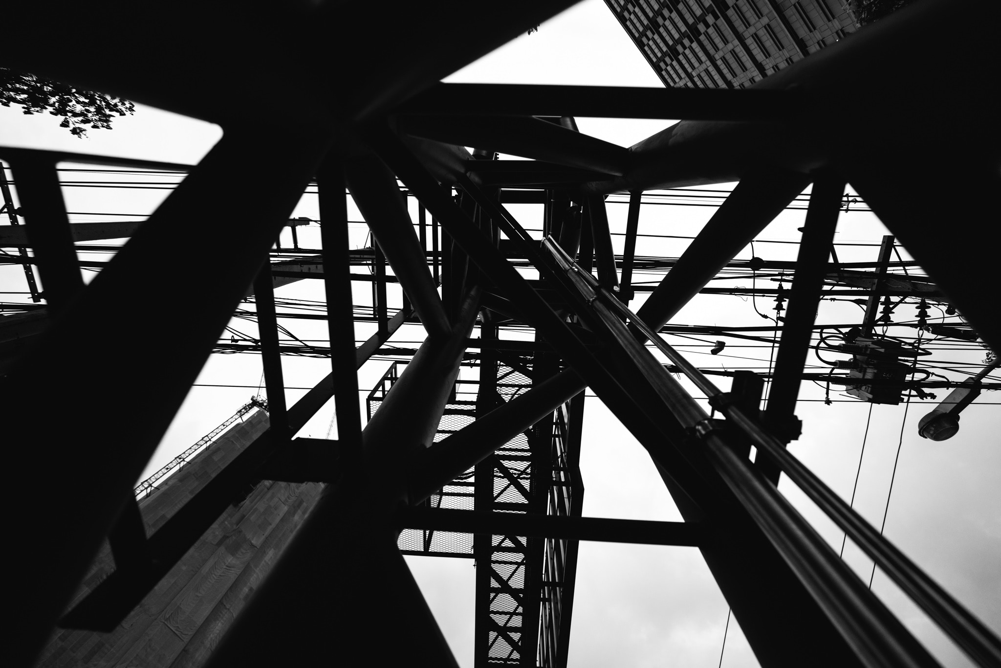 Low-angle black and white view of a metal structure with wires and a building in the background.