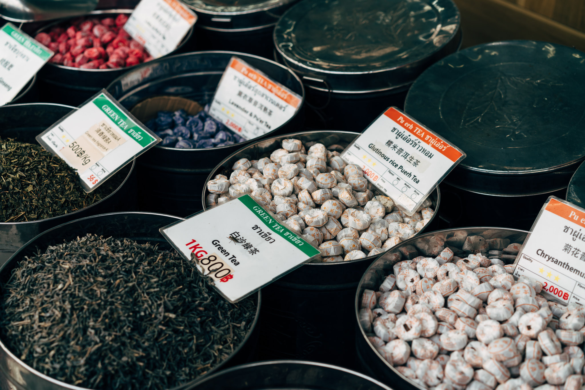 Assortment of loose leaf teas and pressed tea cakes in metal tins with price tags.