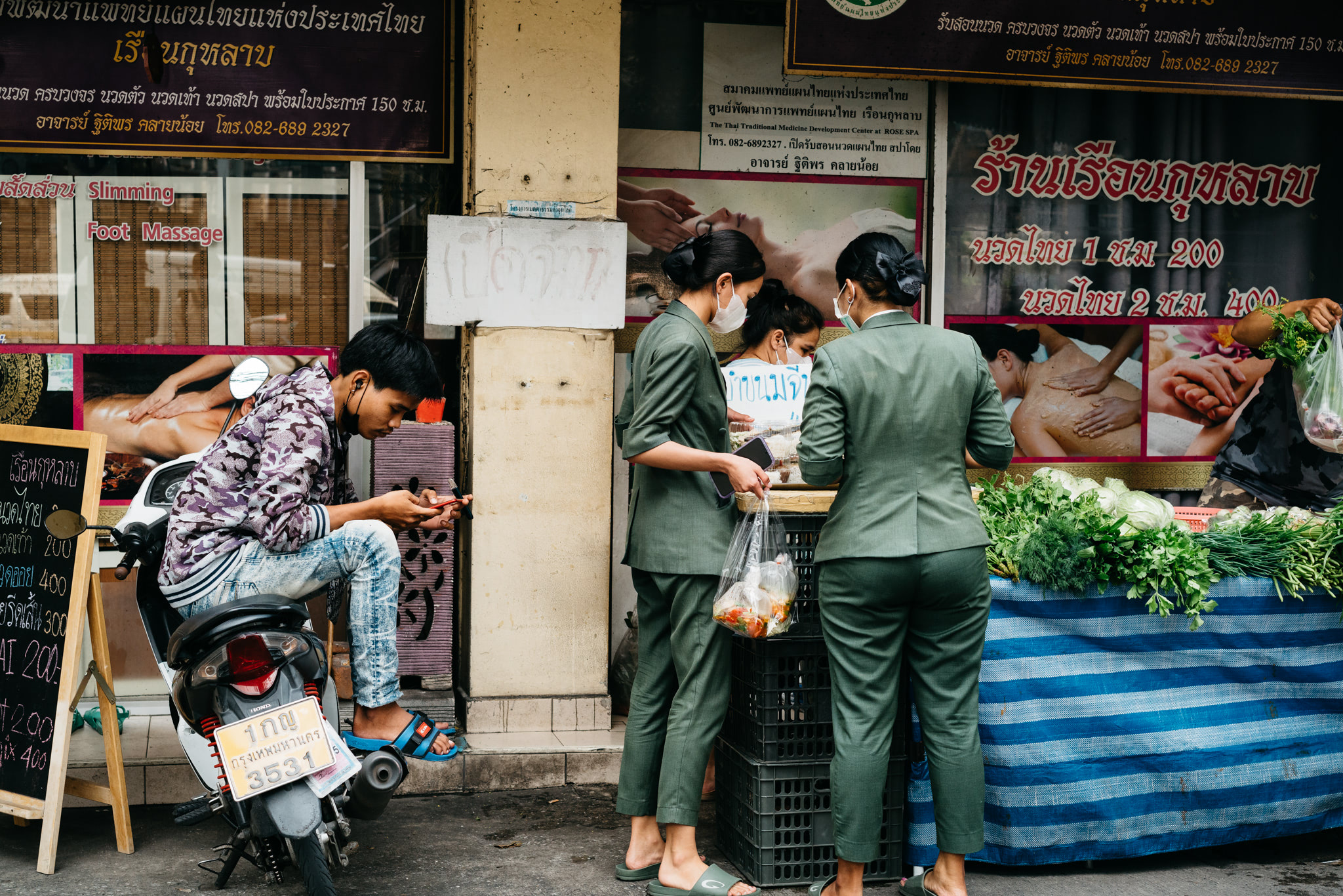 Three women in green uniforms purchase food from a street vendor in Thailand.
