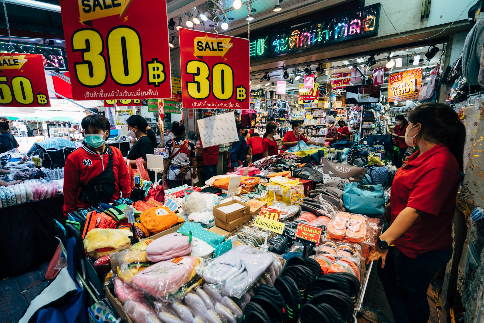 Busy Thai market stall with shoppers wearing masks and various goods for sale, many marked 30฿.