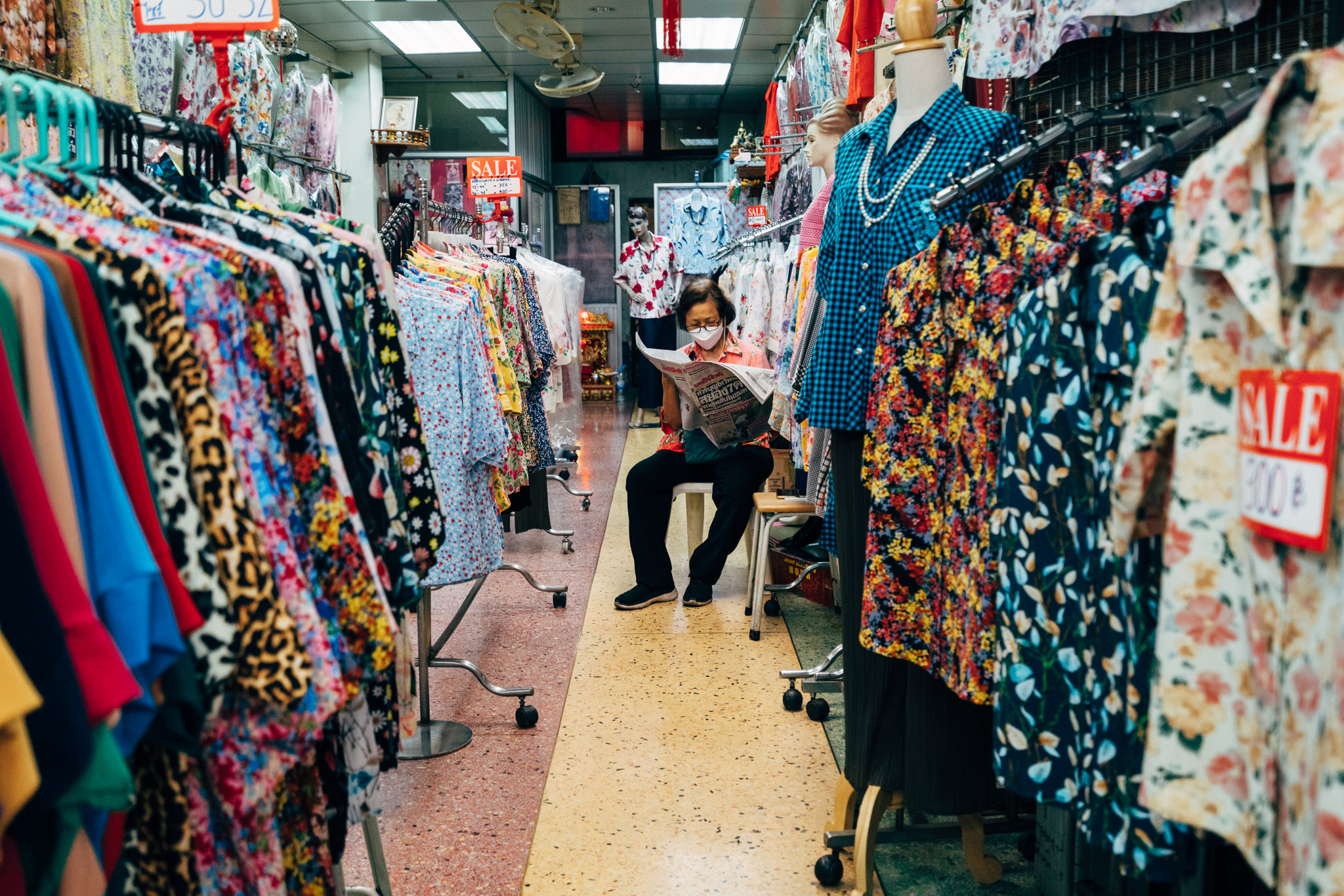 A person wearing a mask sits in a Thai clothing store reading a newspaper; colorful clothes hang on racks on either side.