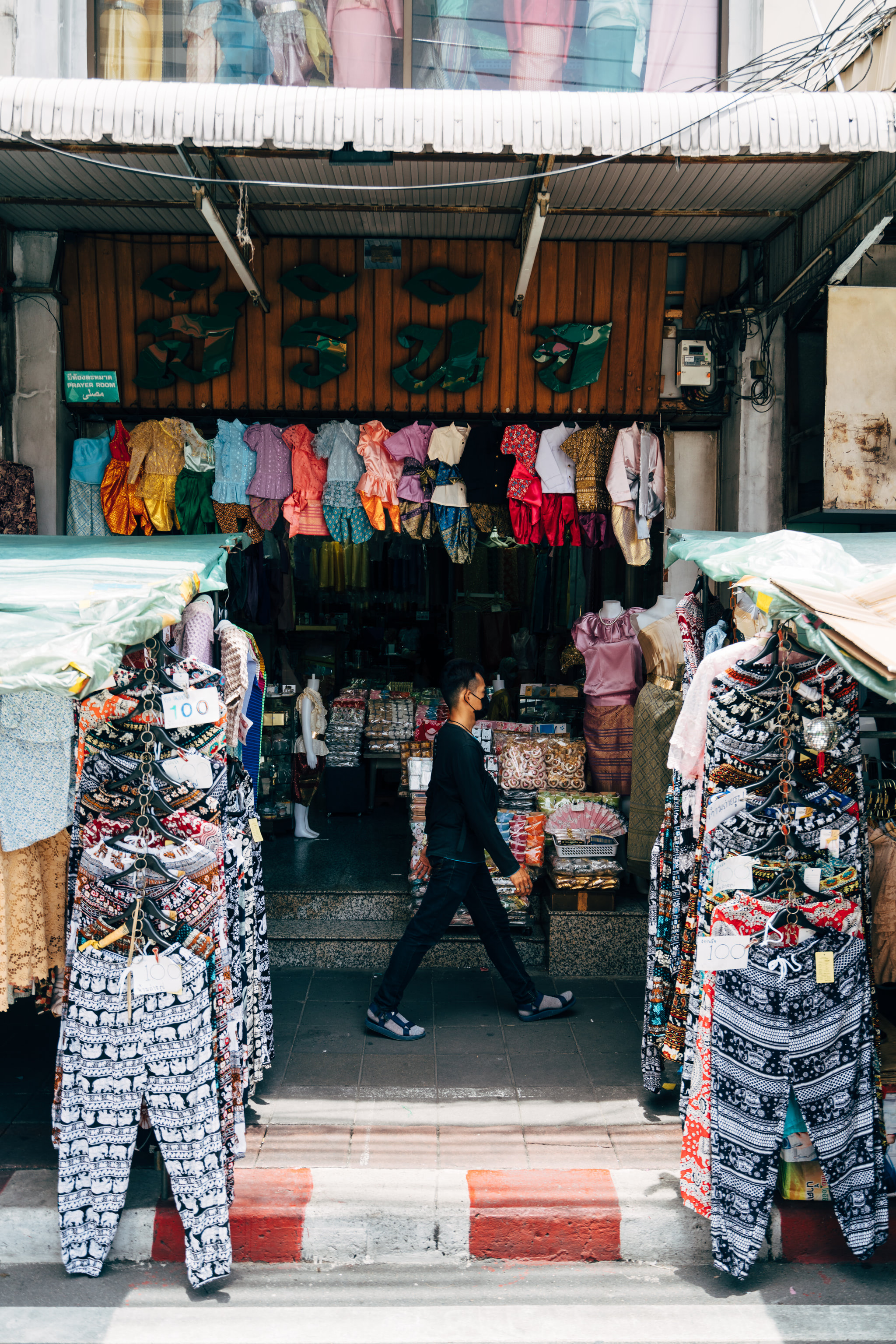 Man walking past a Thai clothing shop with colorful clothing displayed outside.