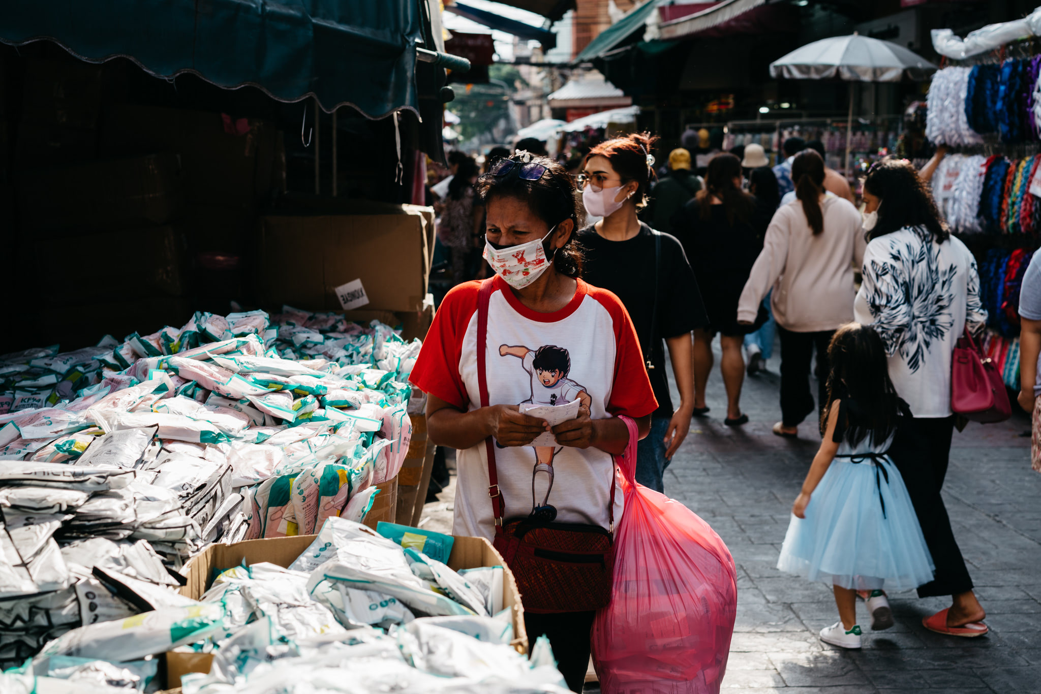 Woman wearing a facemask and red and white shirt looks at a receipt in a busy Thai market.