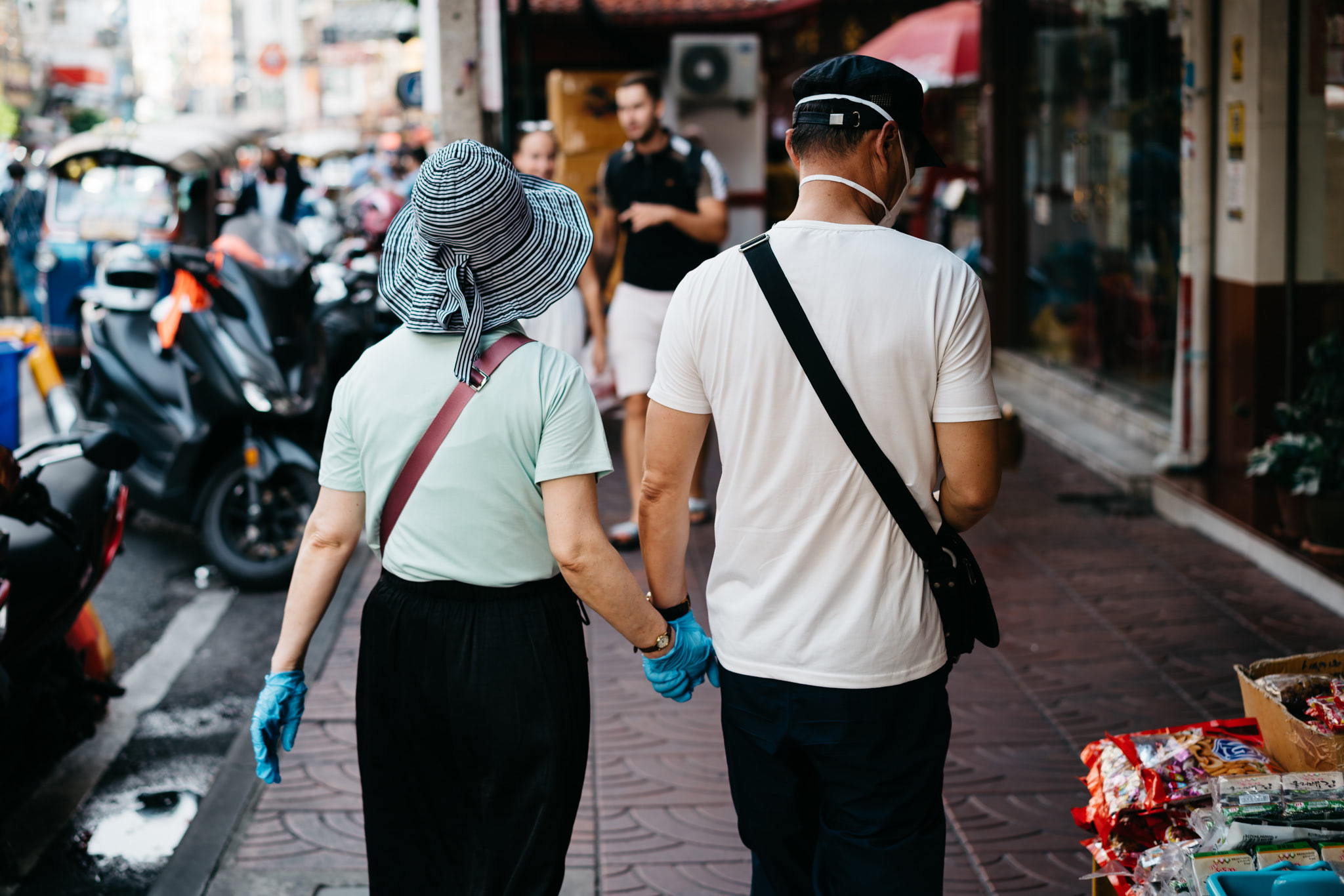 Couple holding hands, wearing face masks and gloves, walking away in a Thai shopping district.