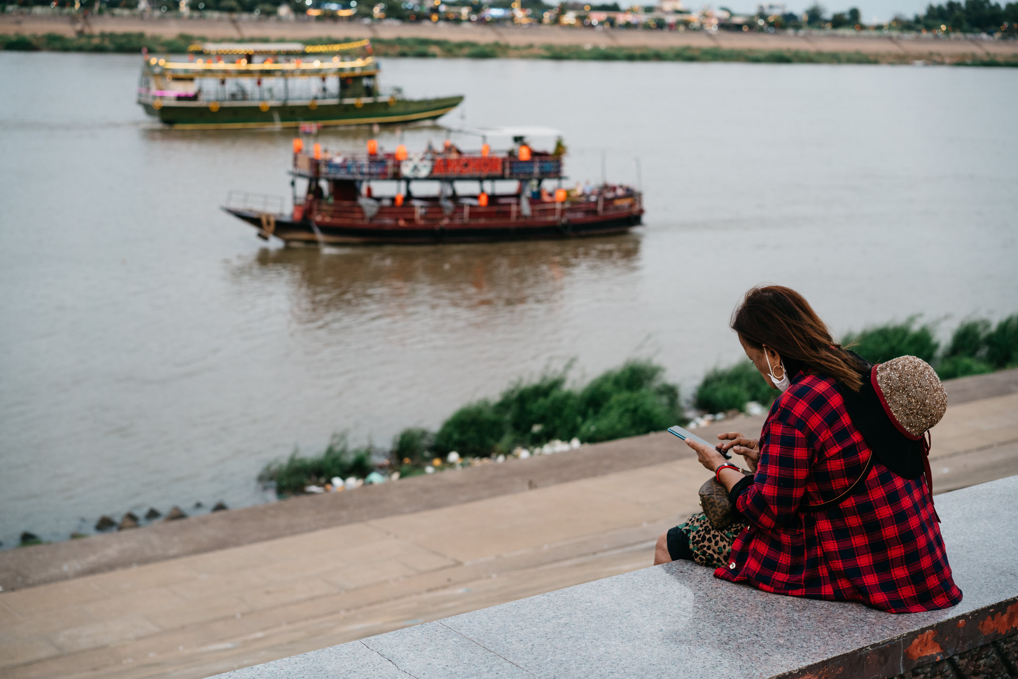 Woman on riverbank in Phnom Penh, Cambodia, using smartphone; two boats on river in background.