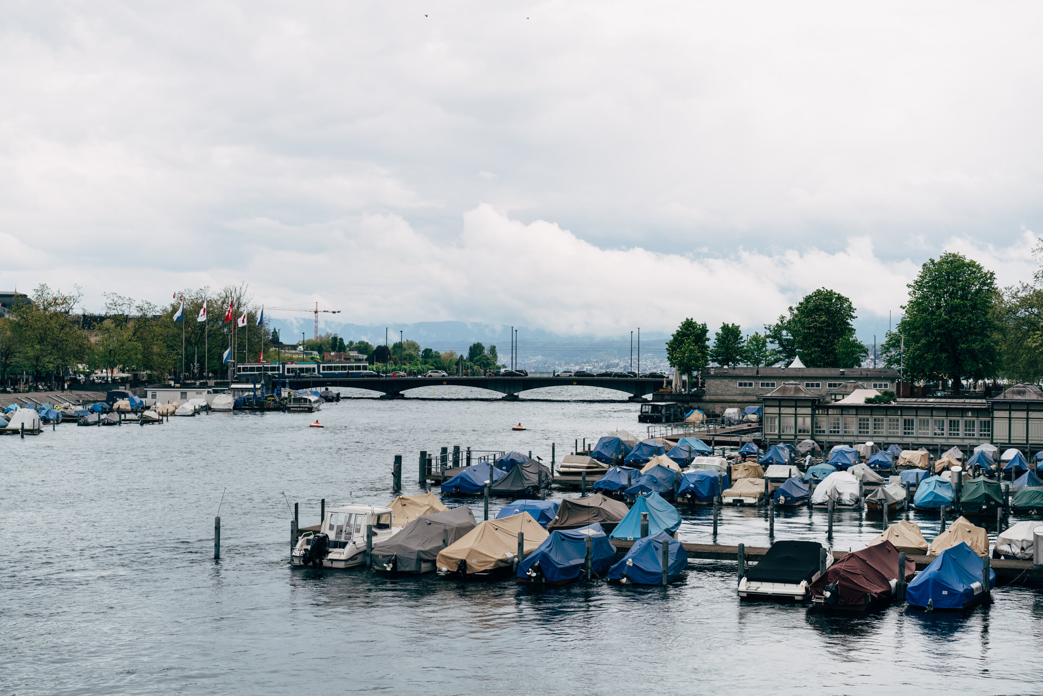 Zurich, Switzerland: Boats covered in tarps at a marina, with a bridge and cityscape in the background.