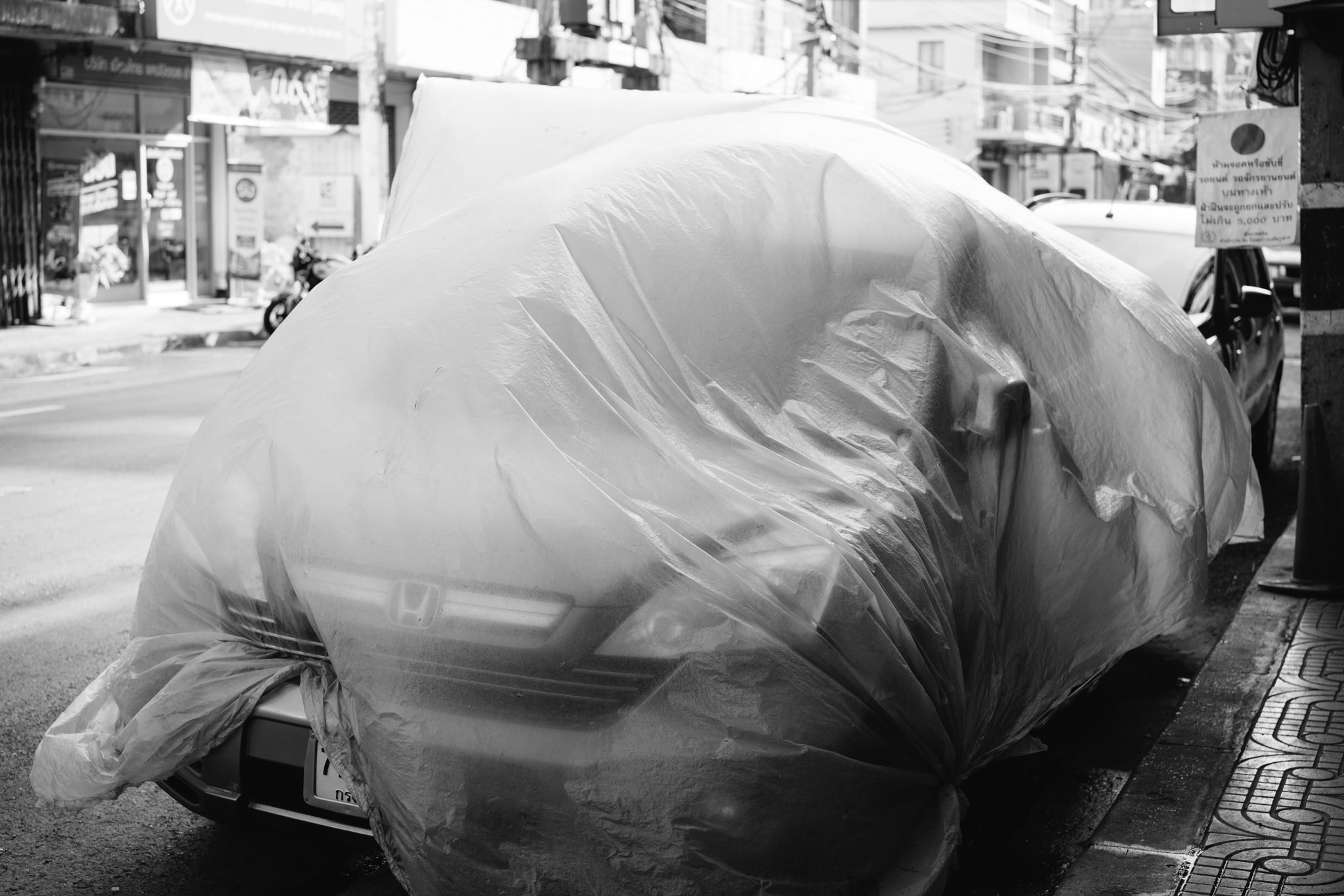 Black and white photo of a car covered in plastic sheeting parked on a city street.