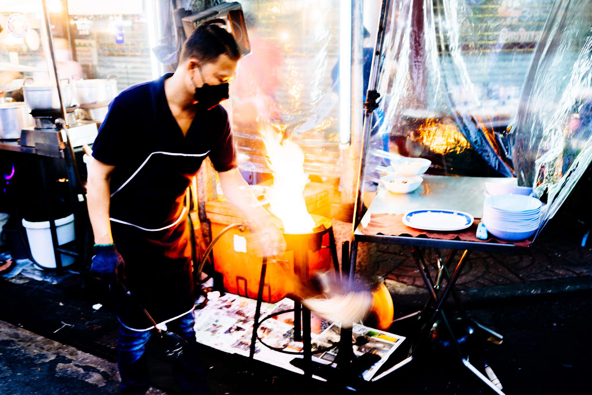 Bangkok street vendor cooking with fire, wearing a mask.