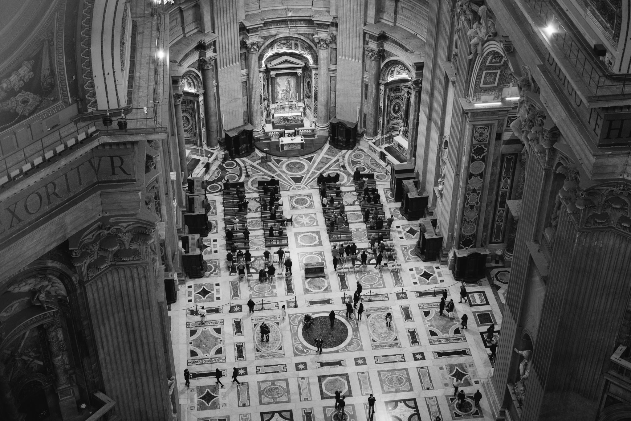 High-angle black and white view of St. Peter's Basilica interior, showing the ornate floor and people sitting in pews.
