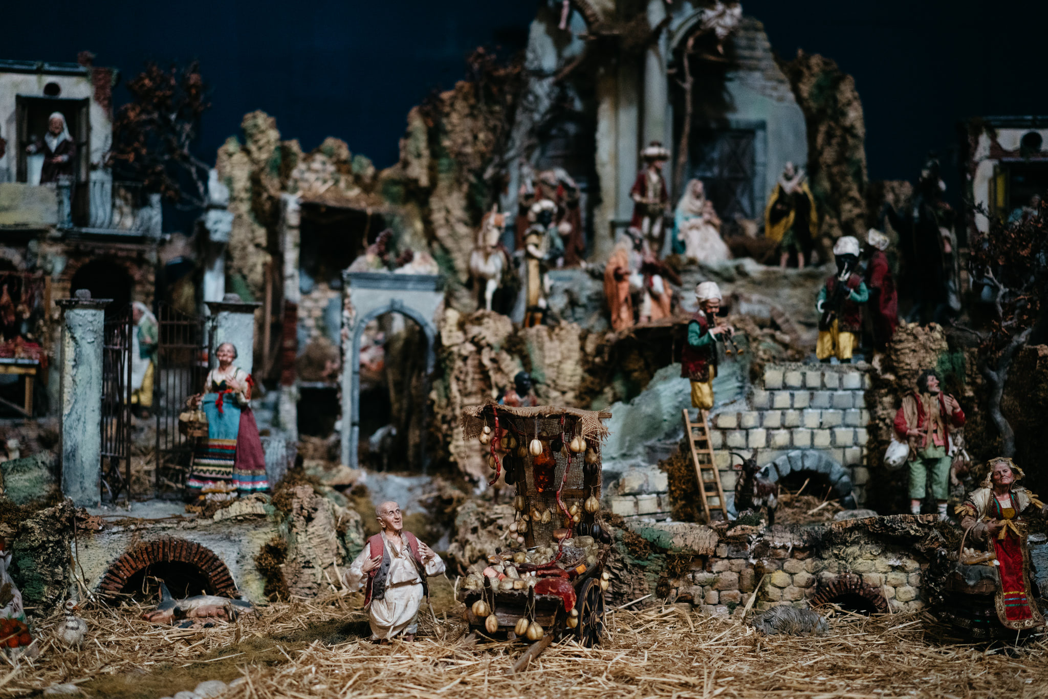 Detailed nativity scene with many figures and a vendor selling produce from a cart.