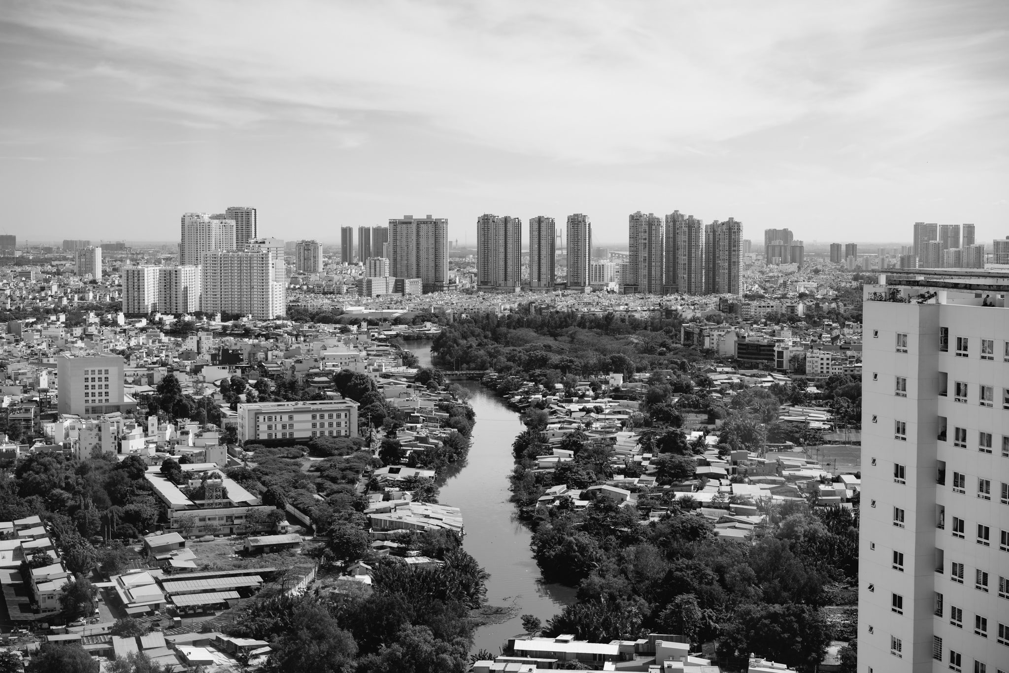 Black and white aerial view of Ho Chi Minh City, showing a river winding through a dense urban landscape of high-rise buildings and low-rise housing.