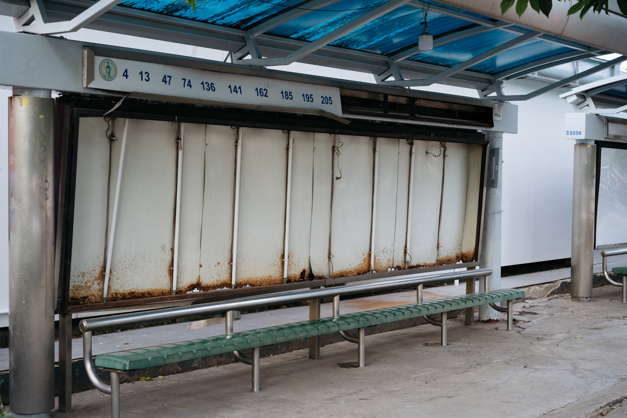 Bus stop with rusty, damaged advertisement panel and green bench; numbers 4, 13, 47, 74, 136, 141, 162, 185, 195, 205 are visible.