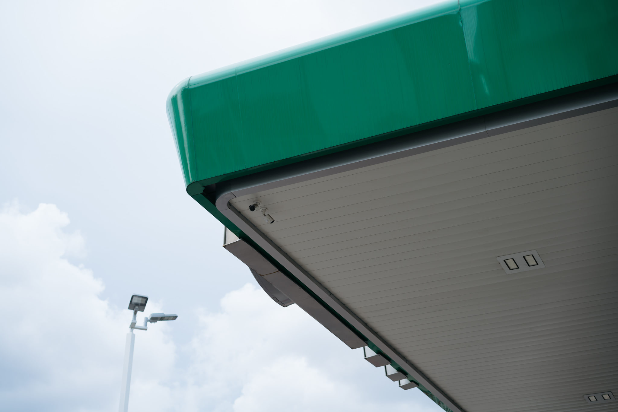 Low-angle view of a gas station canopy with a green top and gray underside, featuring a security camera.