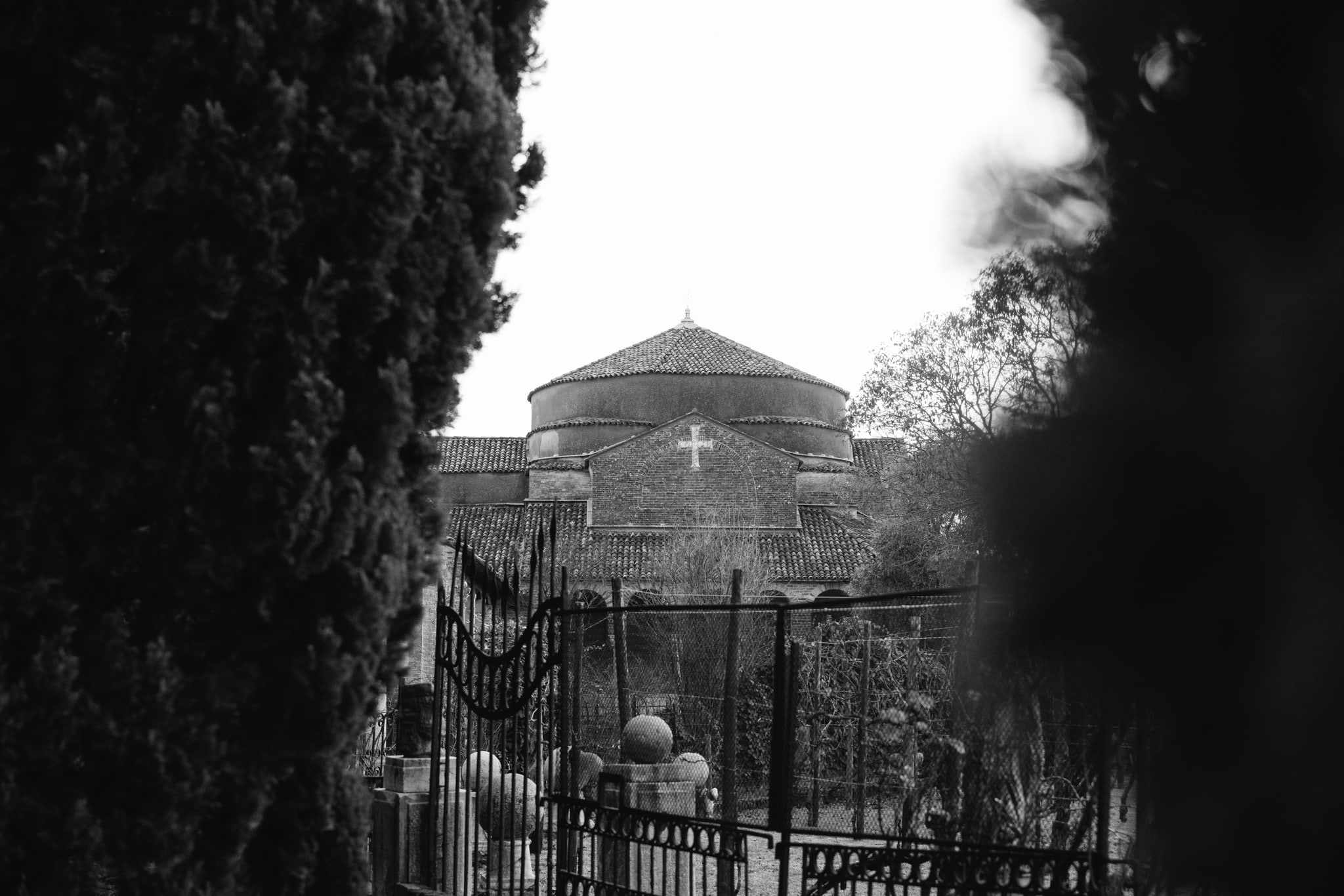 Black and white photo of a church dome in Torcello, Italy, seen through a wrought iron gate and bushes.