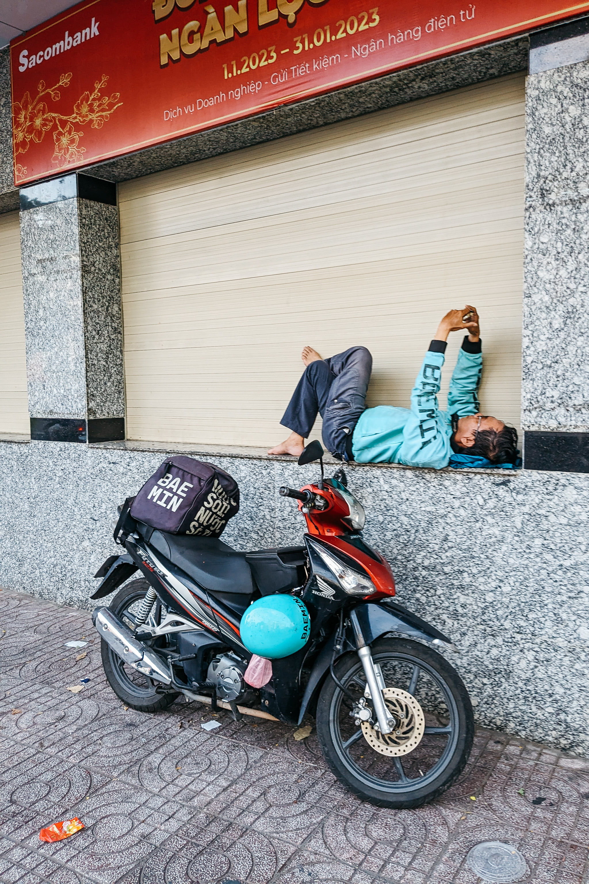 A food delivery driver resting against a wall, looking at his phone, with his motorbike parked nearby.