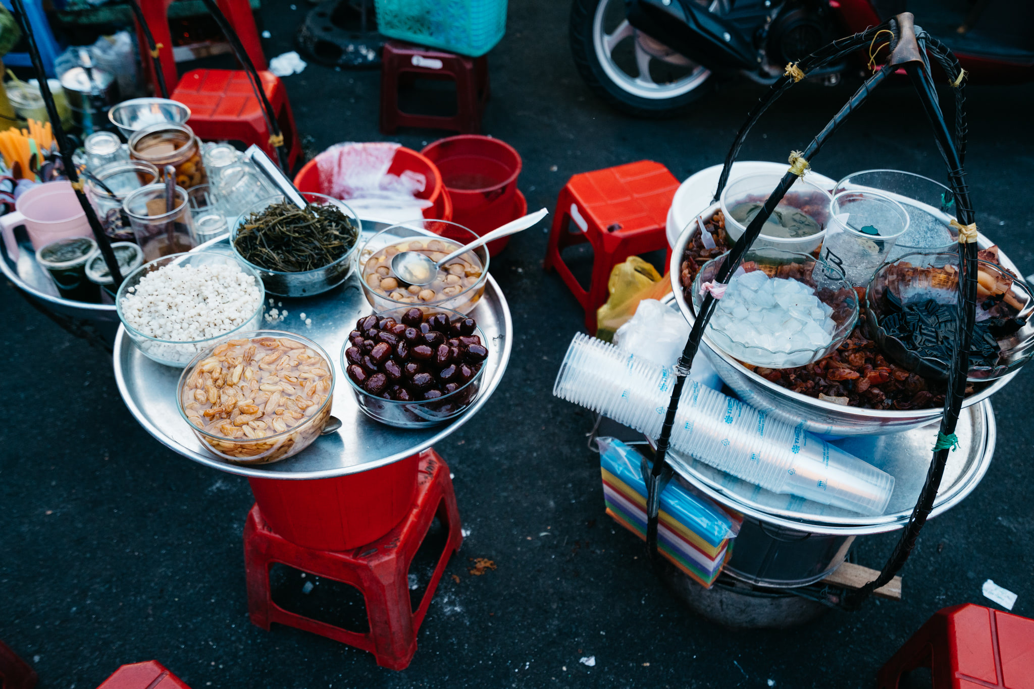 Overhead view of two trays of Vietnamese street food desserts, including various sweet ingredients in bowls.