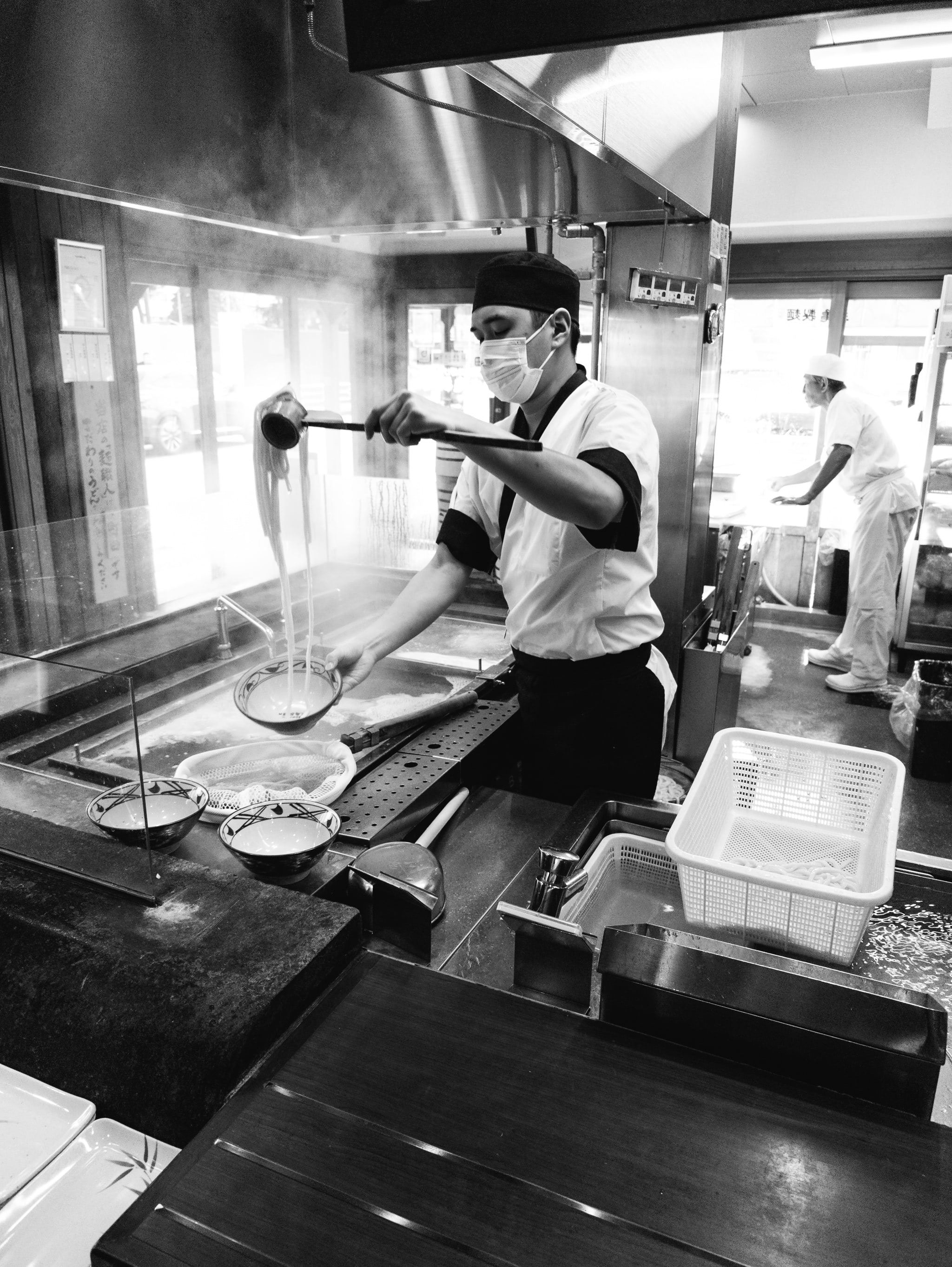 Black and white photo of a chef in a commercial kitchen preparing udon noodles.