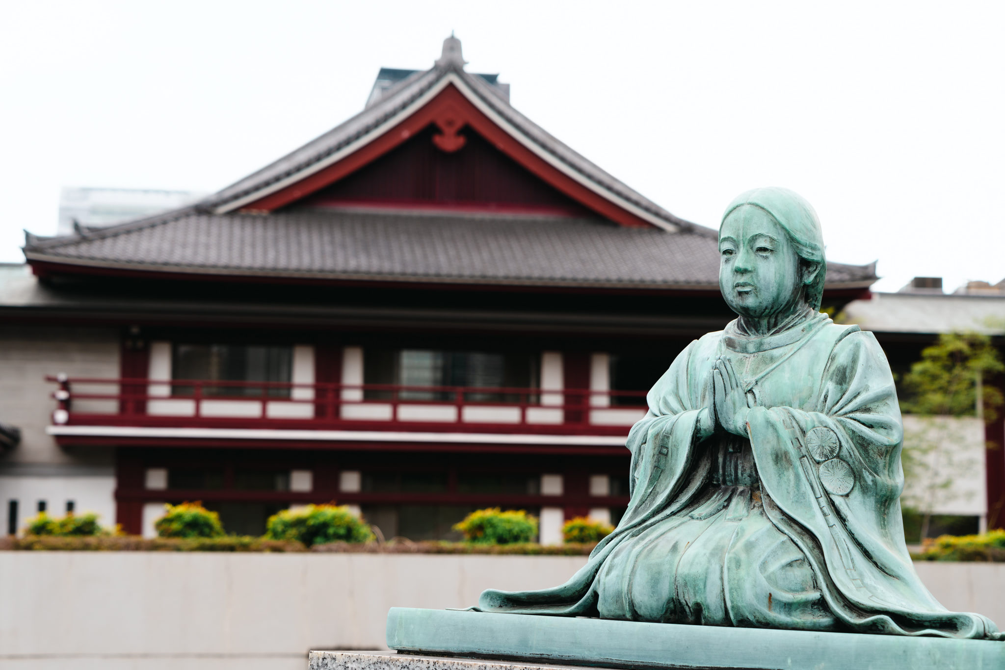 Bronze statue of a person in traditional Japanese clothing praying, in front of a temple.