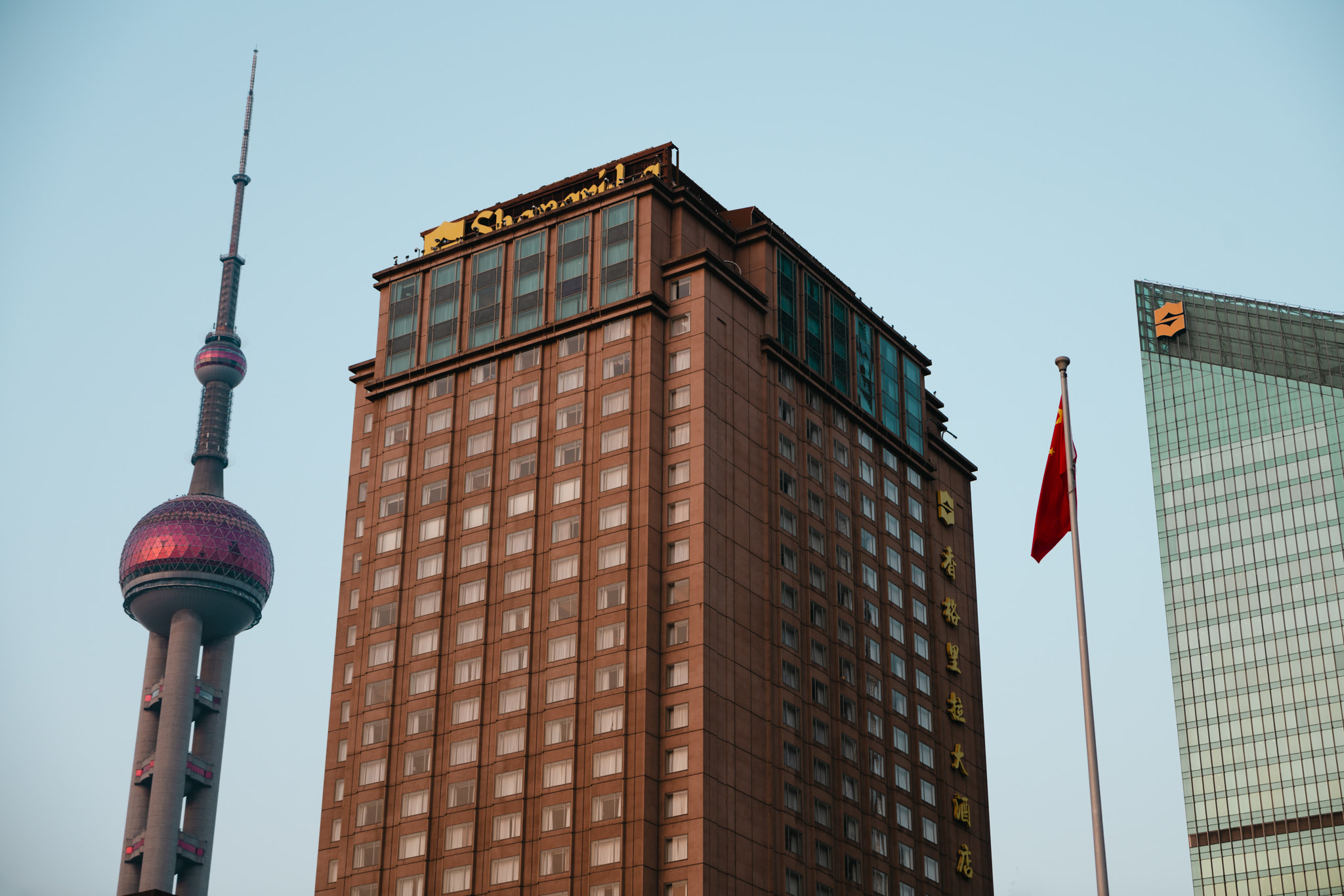 Shanghai Tower, brown hotel building, and Chinese flag against a clear blue sky.