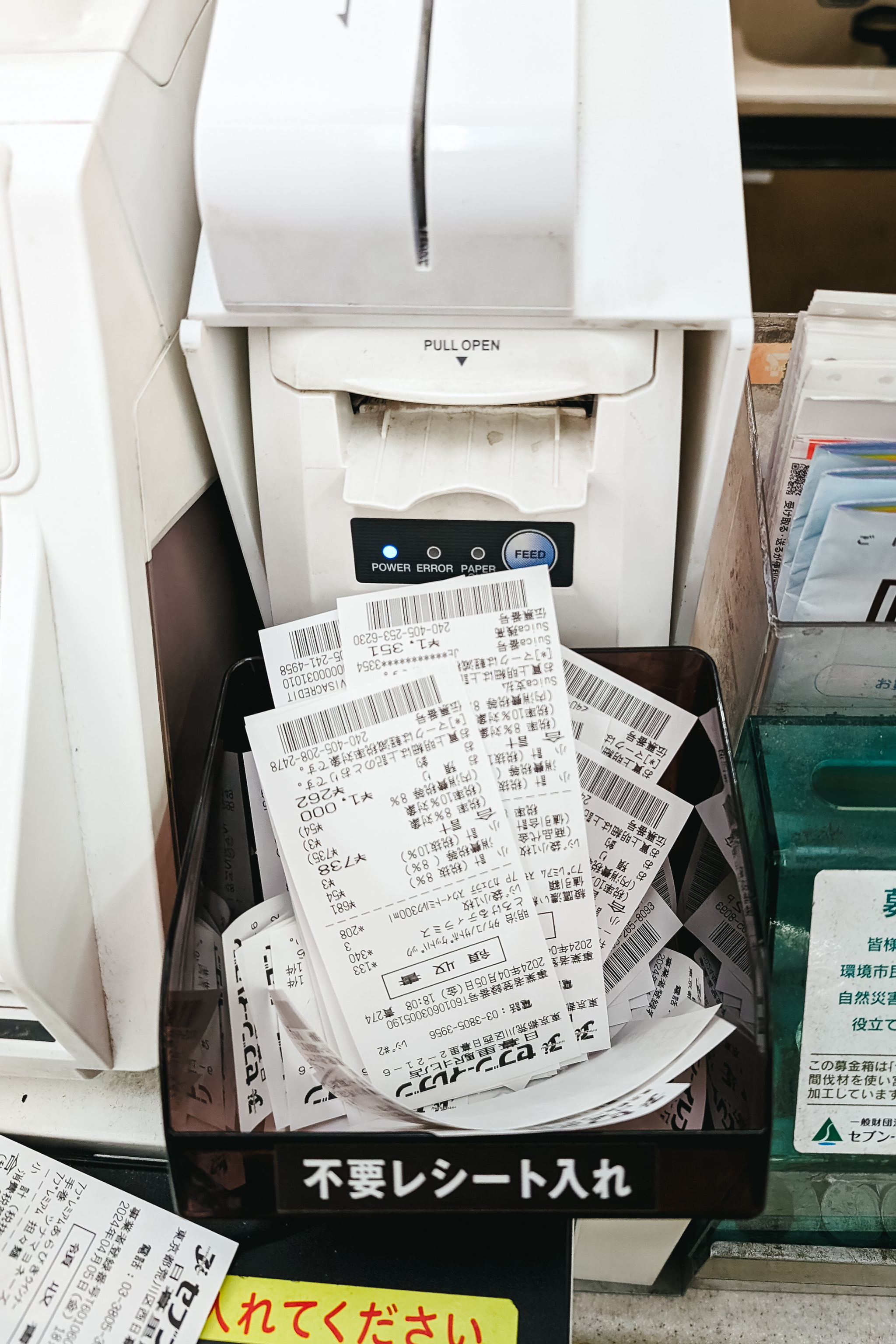 A receptacle labeled 'Unneeded receipts' in Japanese, overflowing with discarded Japanese receipts, sits next to a receipt printer.