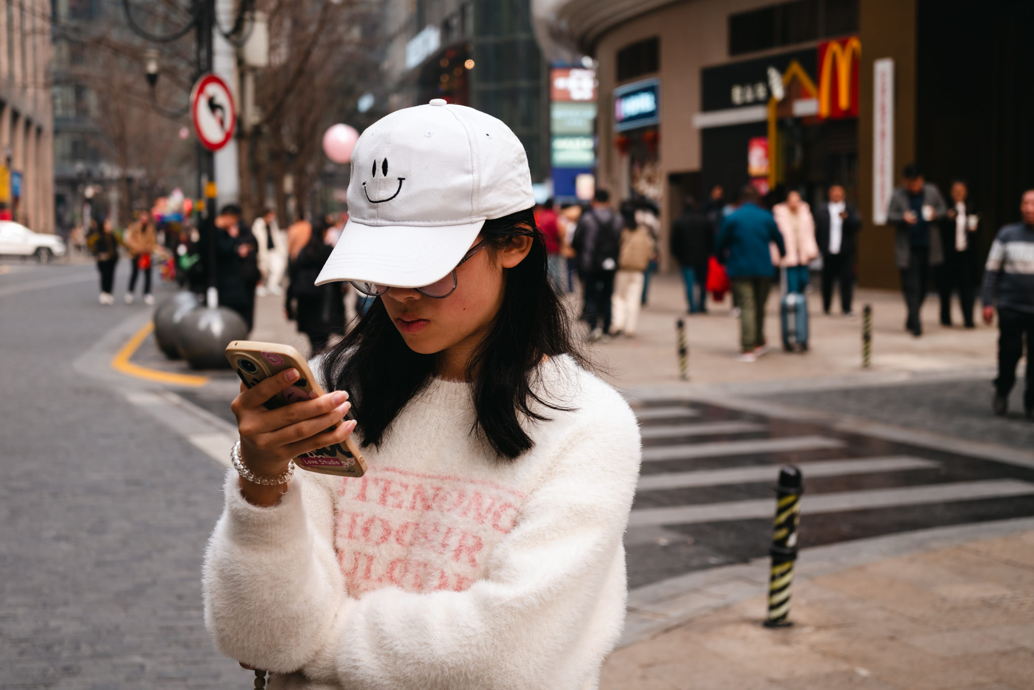 Young woman wearing a white smiley face baseball cap and fluffy sweater uses her phone on a city street.