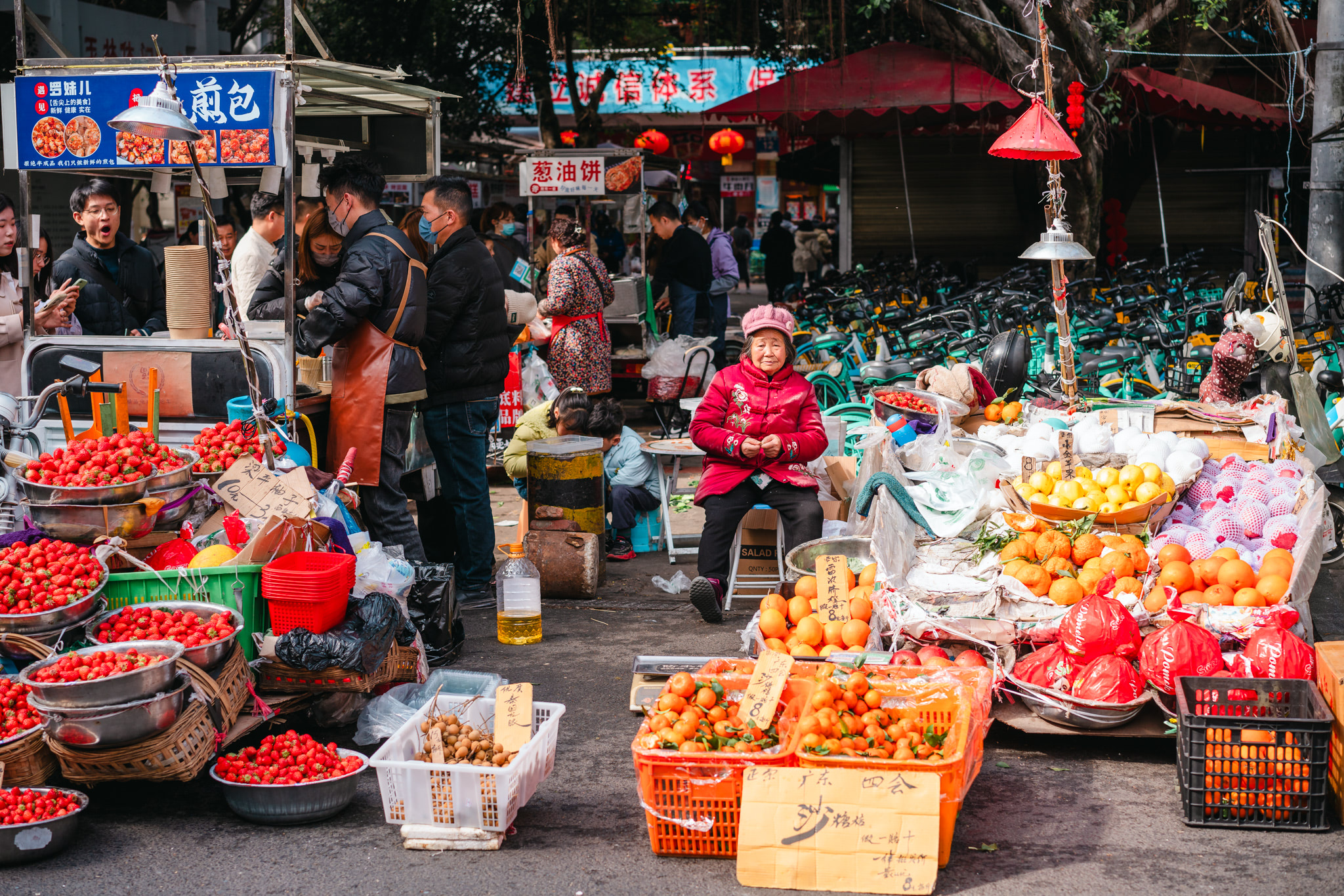A bustling Chengdu street market scene with vendors selling fruit, including strawberries, oranges, and mandarins, and a woman in a red jacket sitting at a stall.