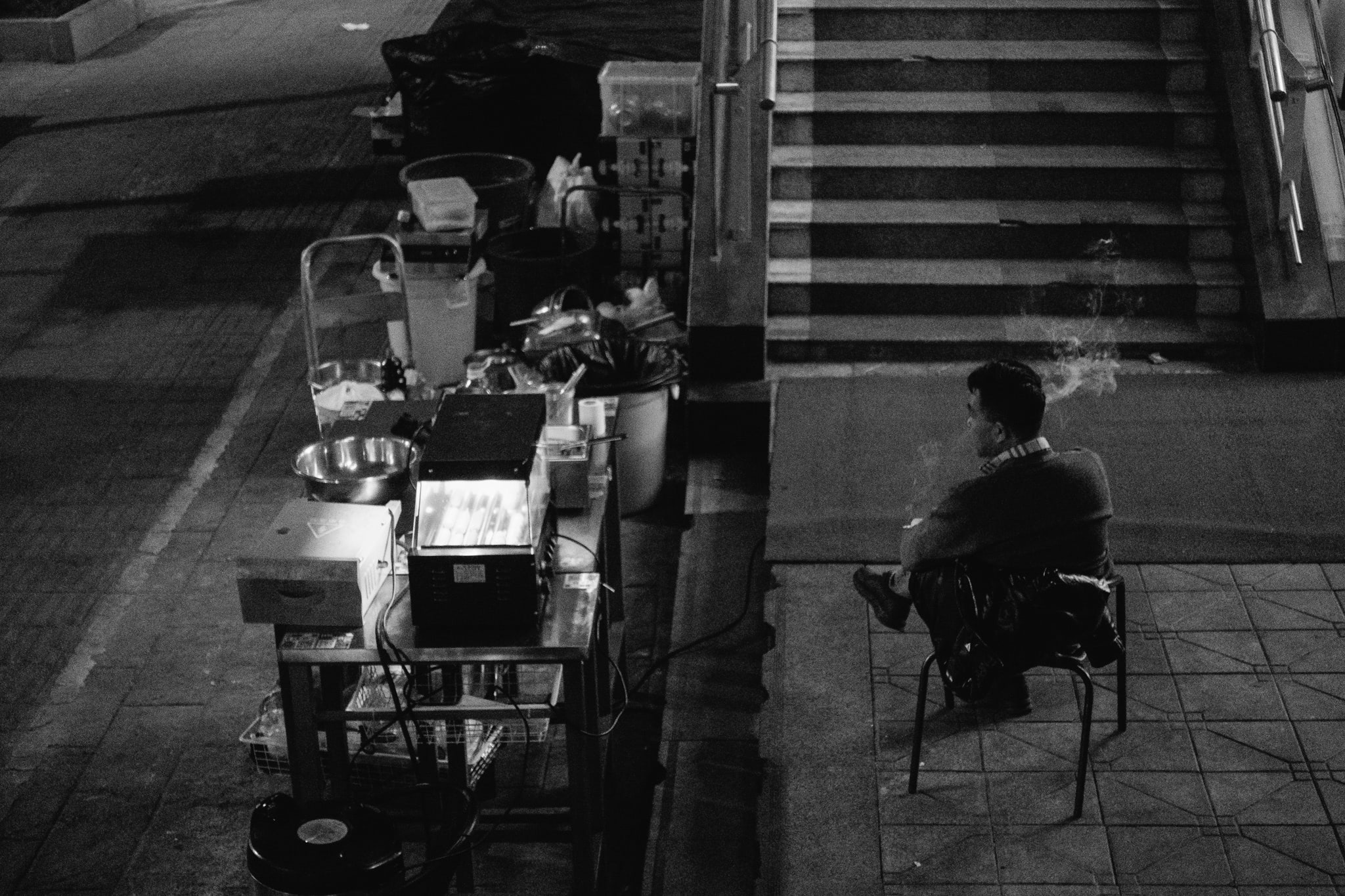 Black and white photo of a man smoking, sitting on a chair next to a food cart at night.