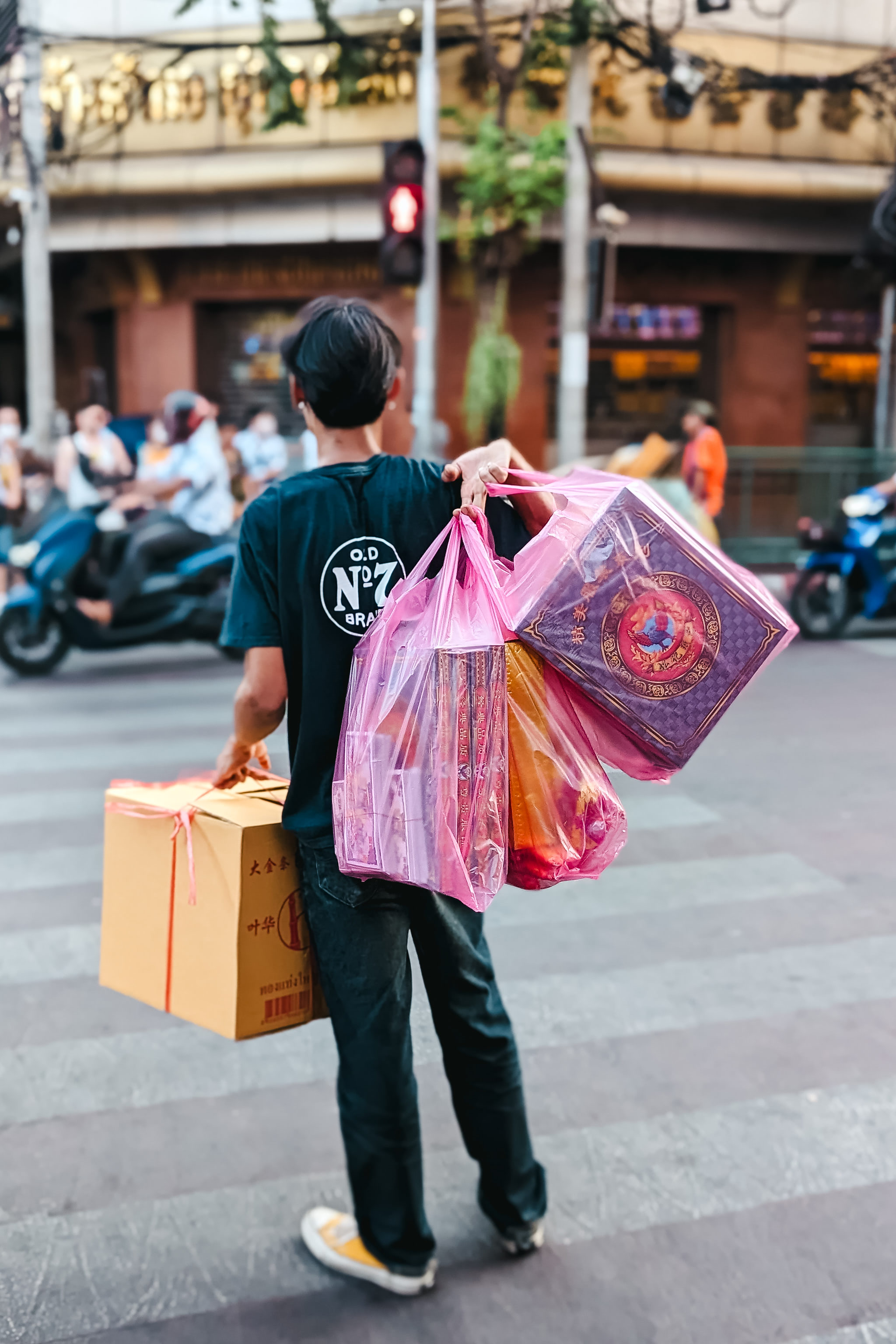 Person carrying cardboard box and plastic bags filled with items on a Bangkok street.