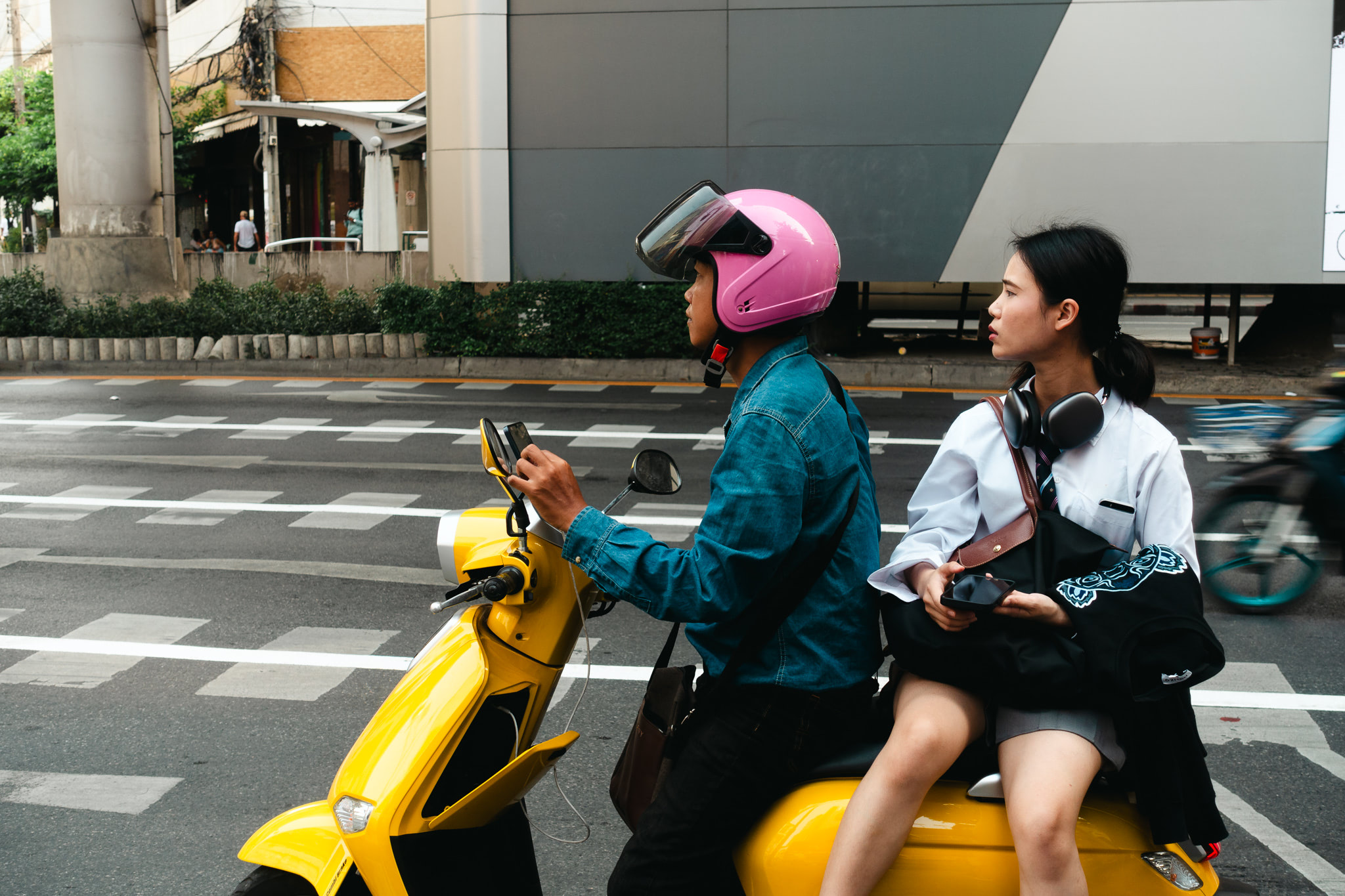A yellow motorbike taxi in Bangkok, driven by a man wearing a pink helmet, with a woman passenger.