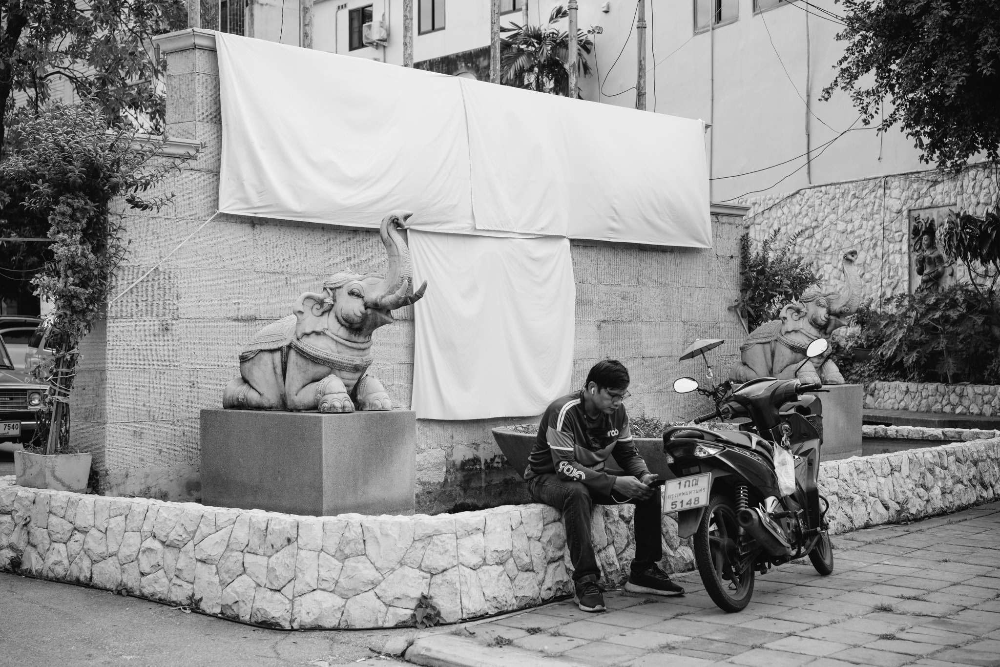 Black and white photo of a man sitting on a low wall next to a motorbike, with elephant statues on either side.