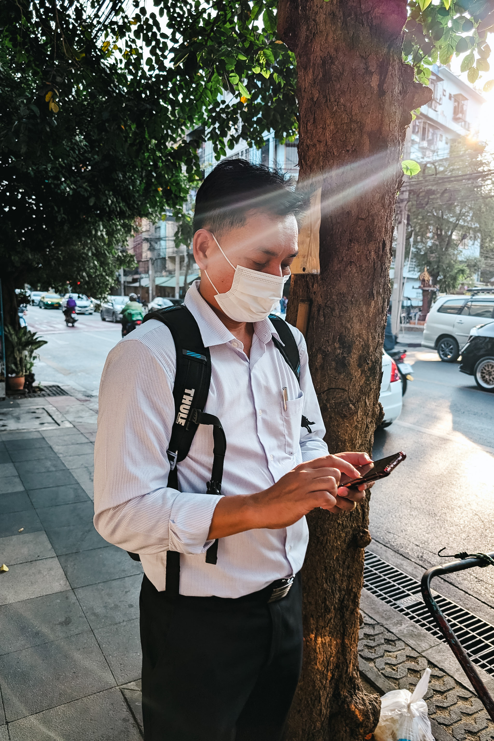 Man wearing a mask and backpack checks his phone on a city sidewalk.