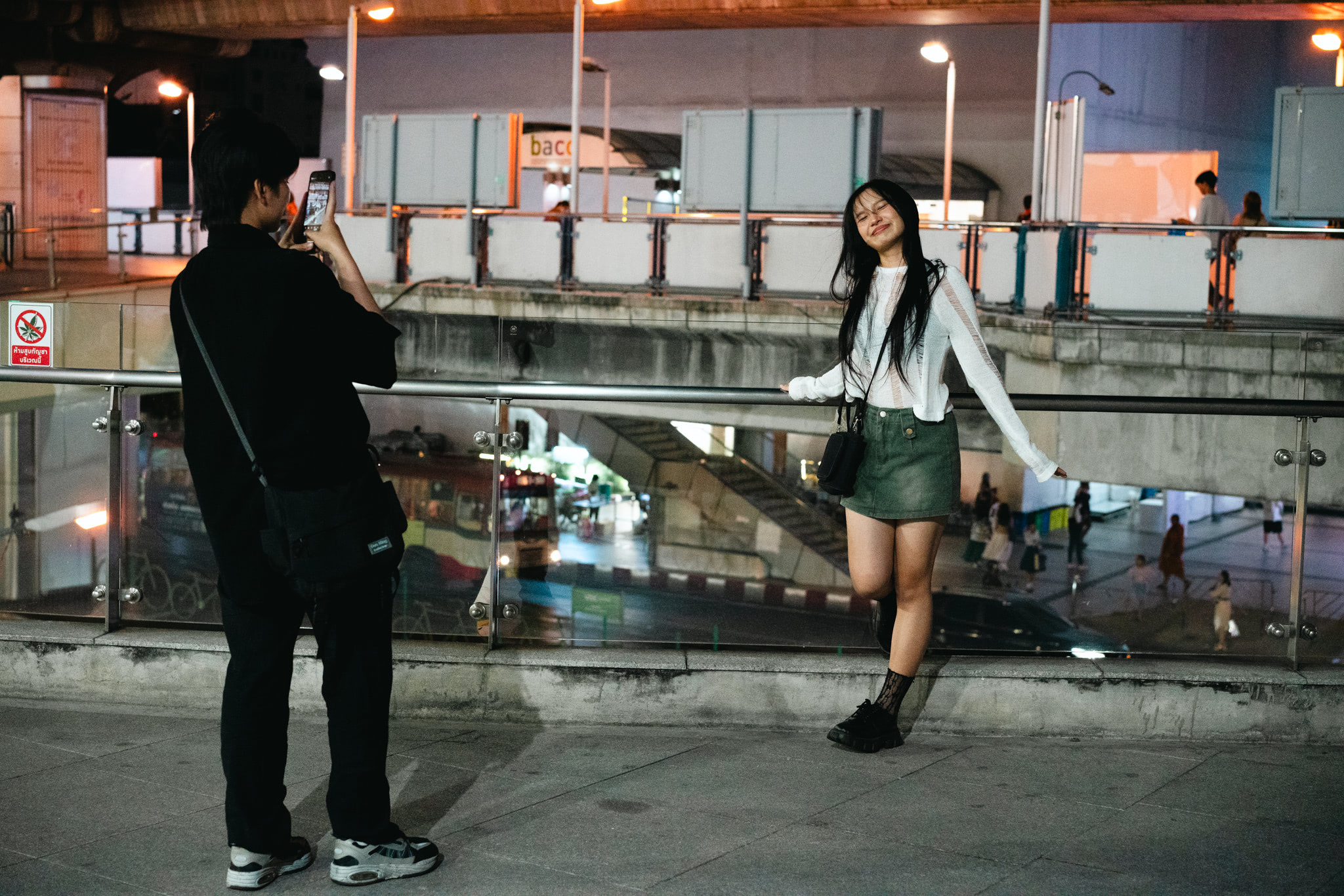 A person photographs a woman leaning on a railing at night in Bangkok.