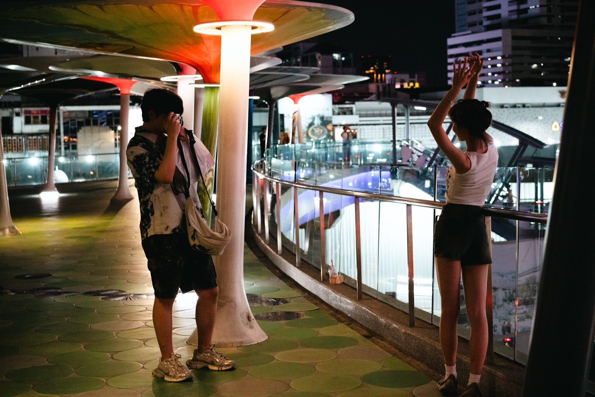 A person photographs a woman posing at night on a walkway with illuminated mushroom-shaped structures.