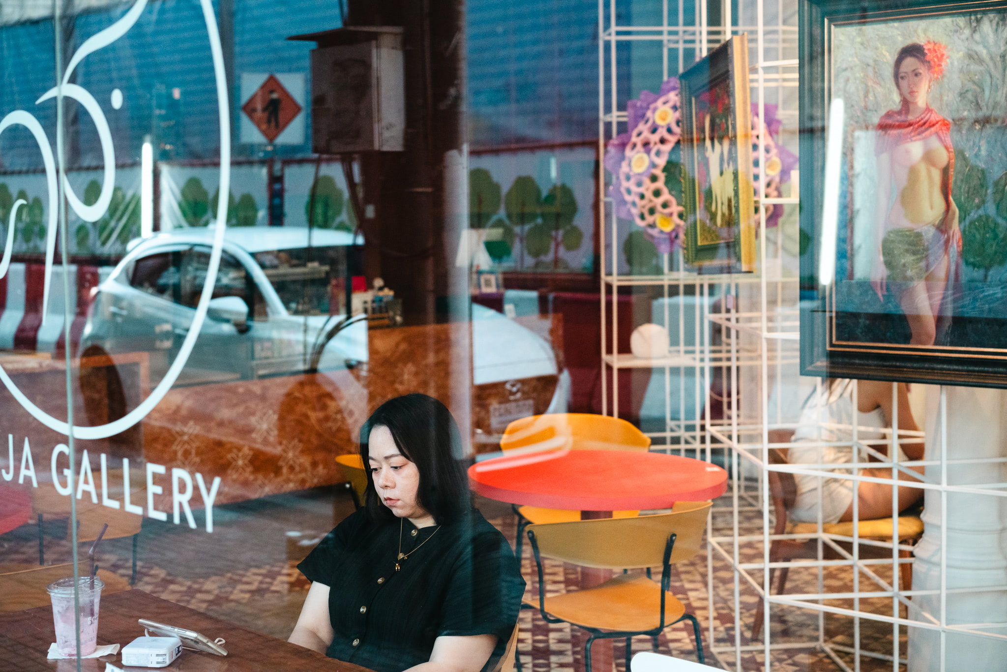 Woman sitting at a cafe table looking at a phone, viewed through a gallery window.