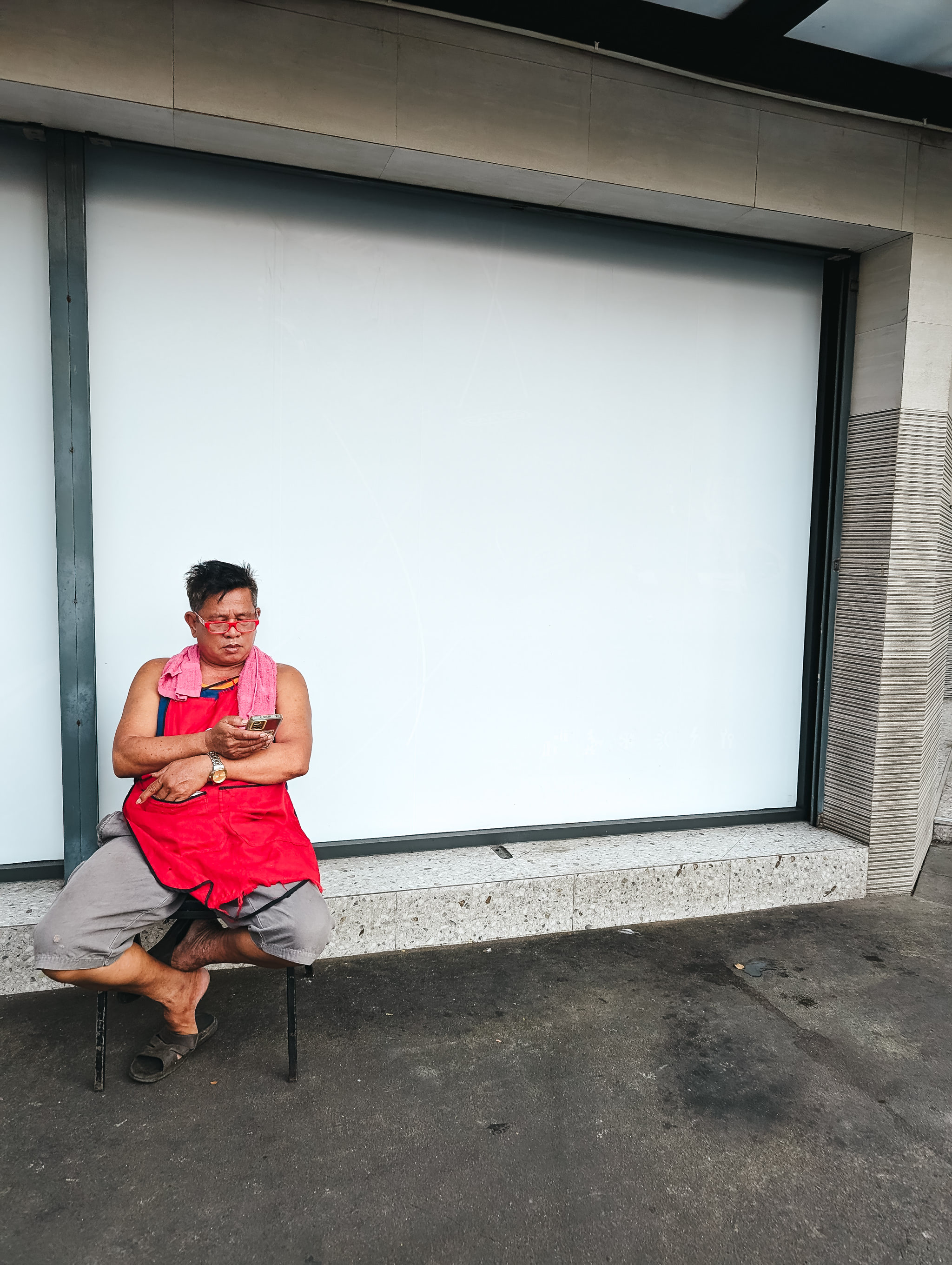Man in red apron sitting on chair outdoors, looking at phone.