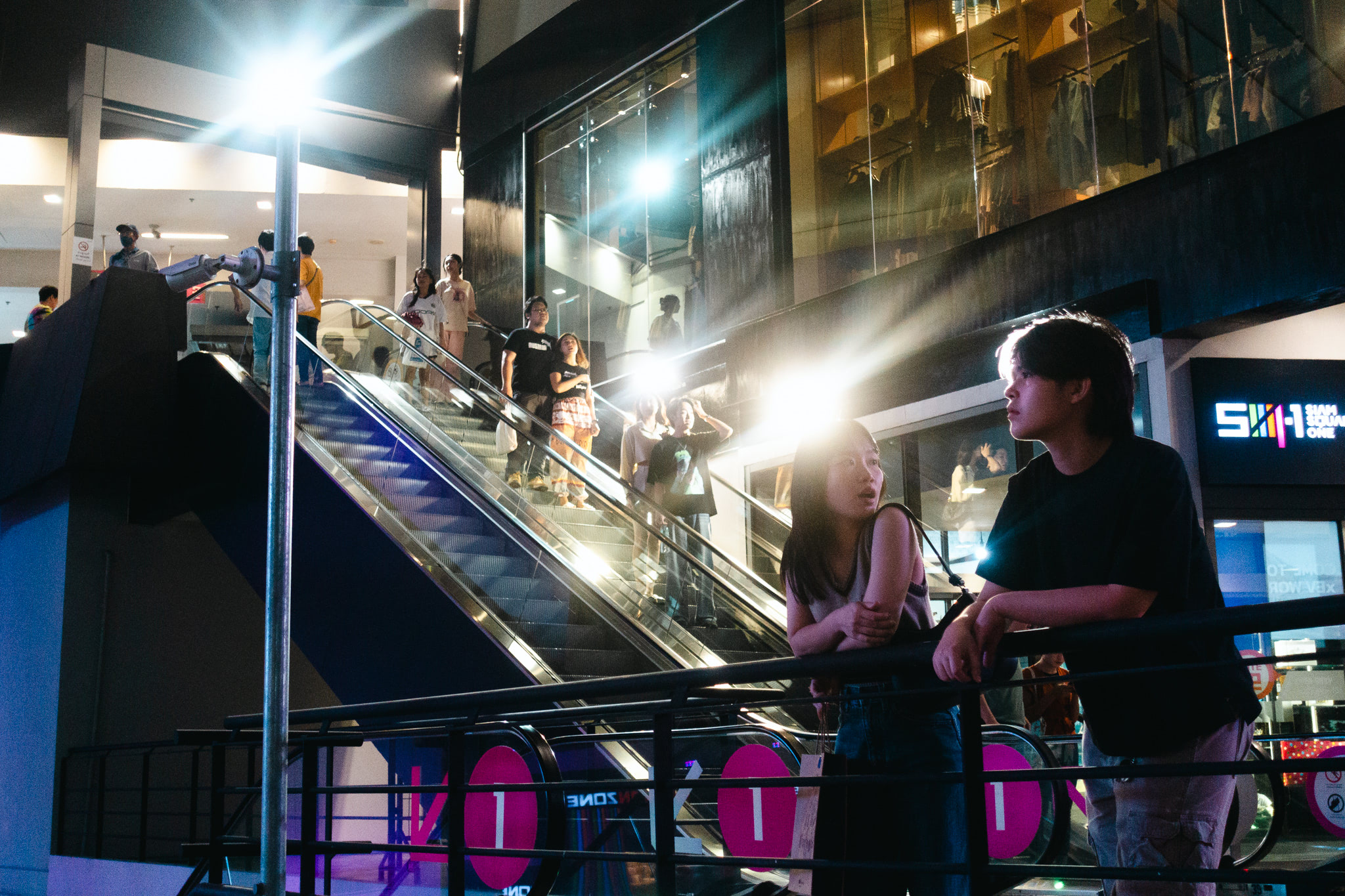 Two young people stand by an escalator in a brightly lit shopping mall at night.