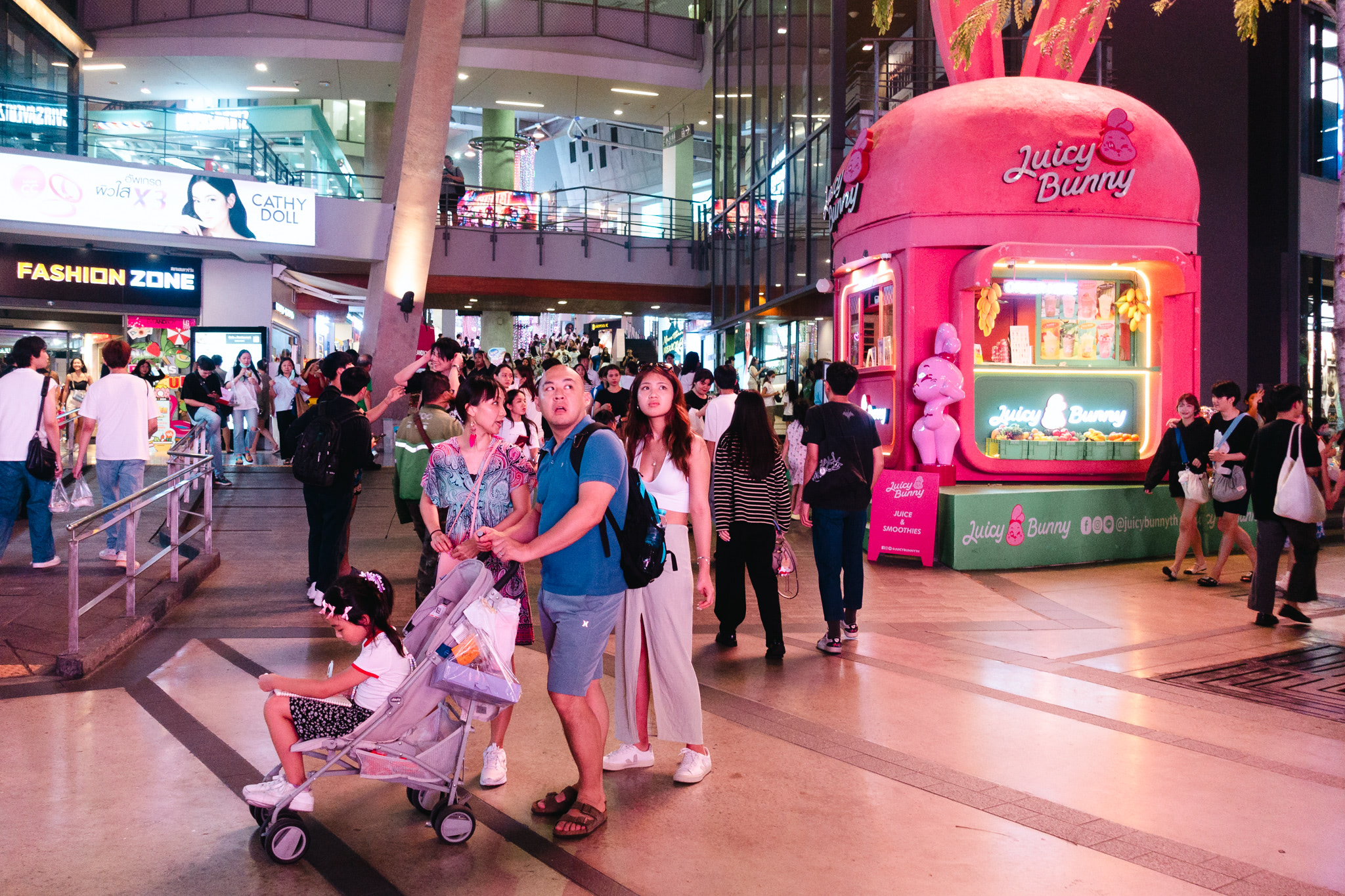 A family with a young child in a stroller walks past a brightly lit Juicy Bunny juice and smoothie stand in a bustling Asian shopping mall.
