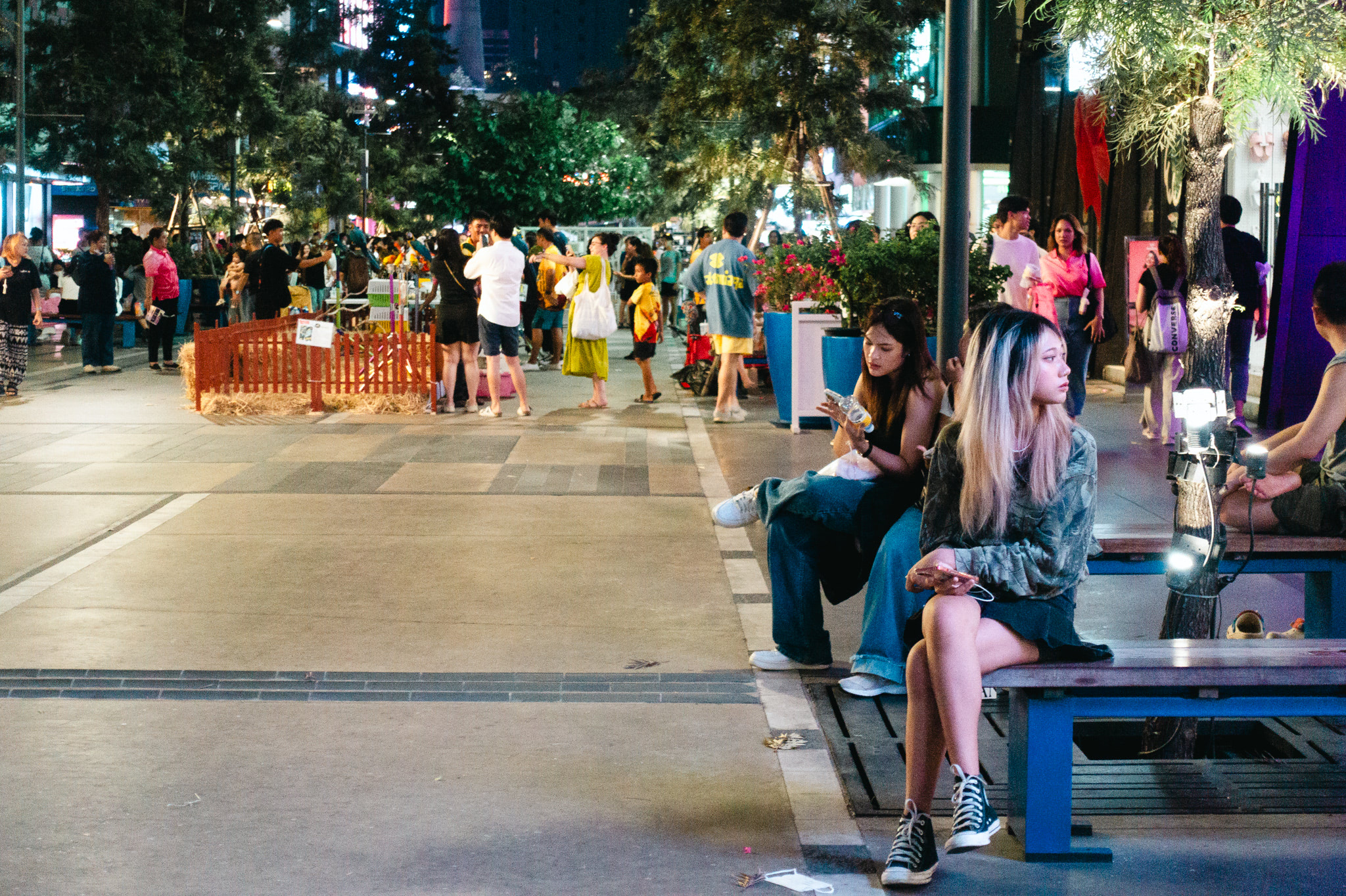 Two young women sit on a bench at night in a crowded outdoor plaza.