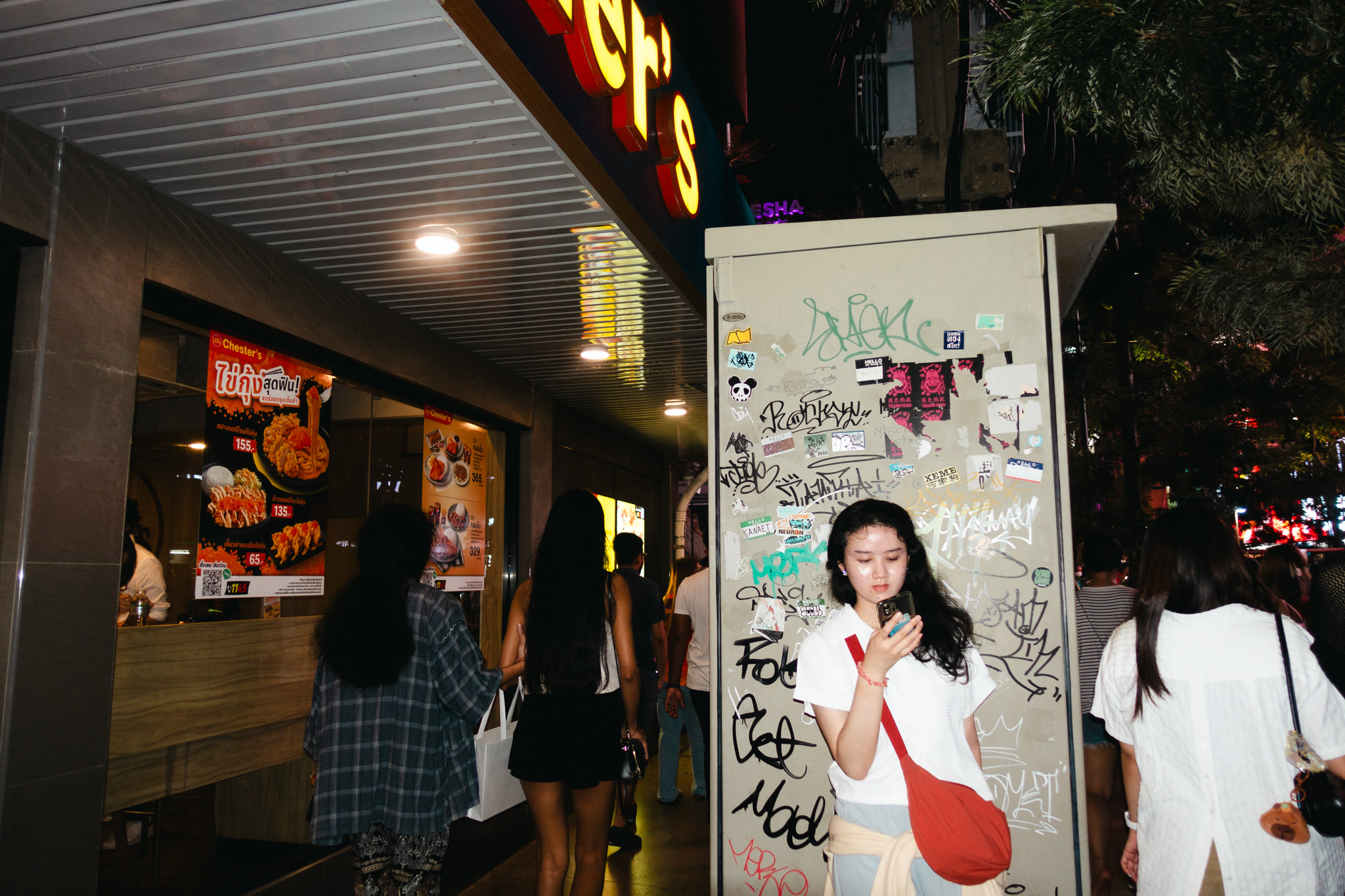 Young woman on phone, graffitied wall, Thai street scene at night.