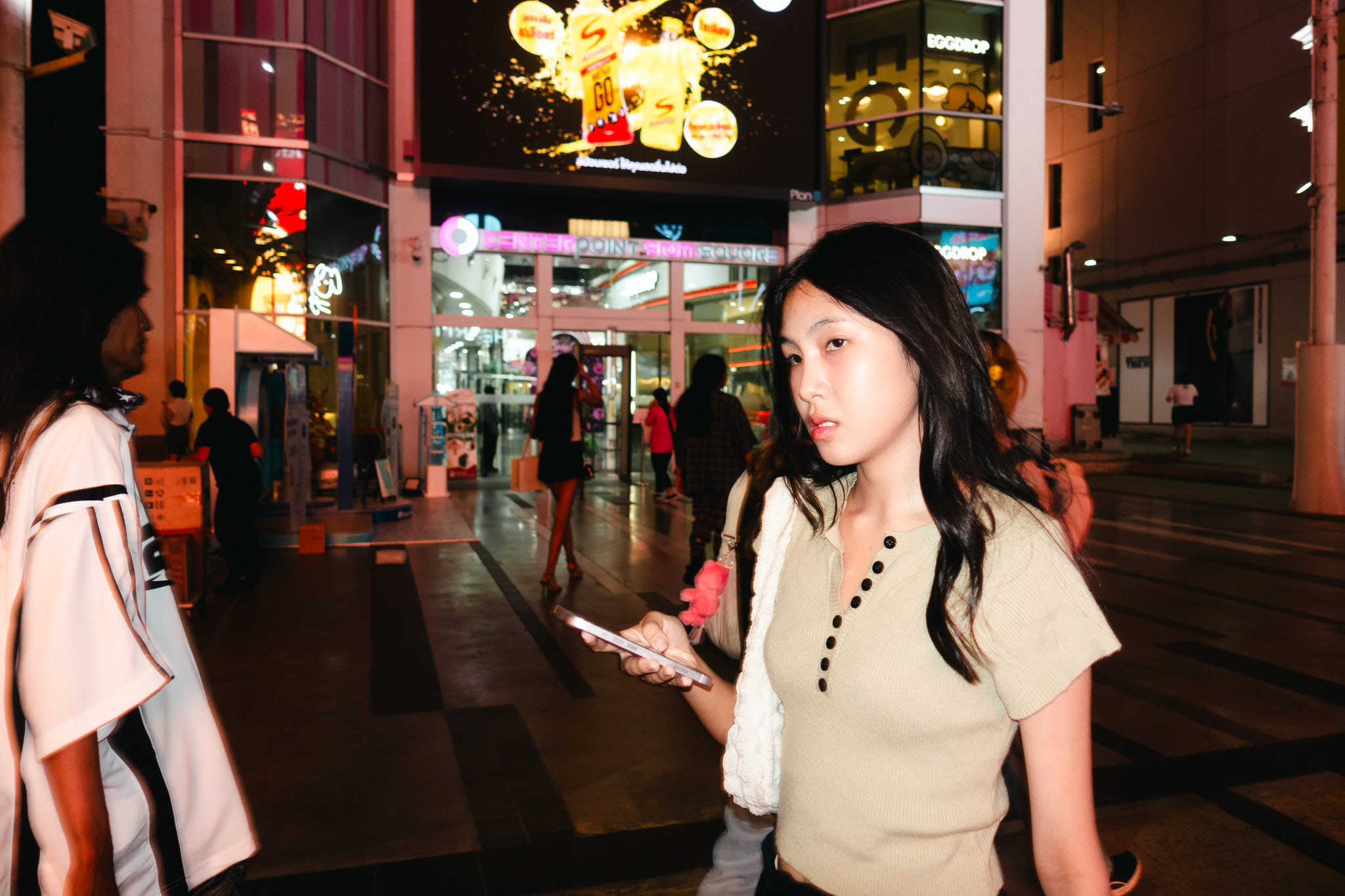 Young woman using smartphone in front of a brightly lit shopping mall at night.