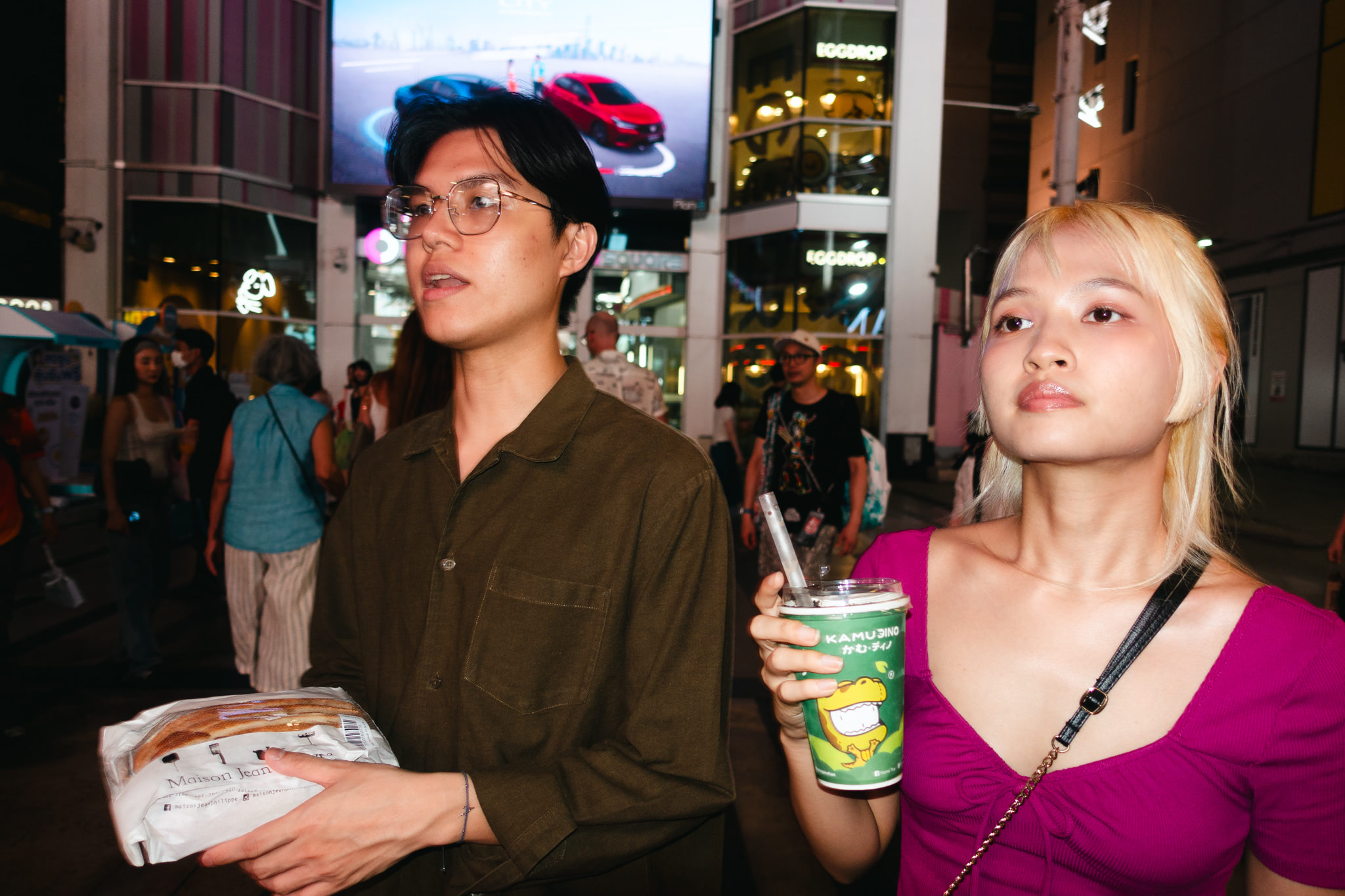 Young man and woman on a city street at night, holding food and a drink.