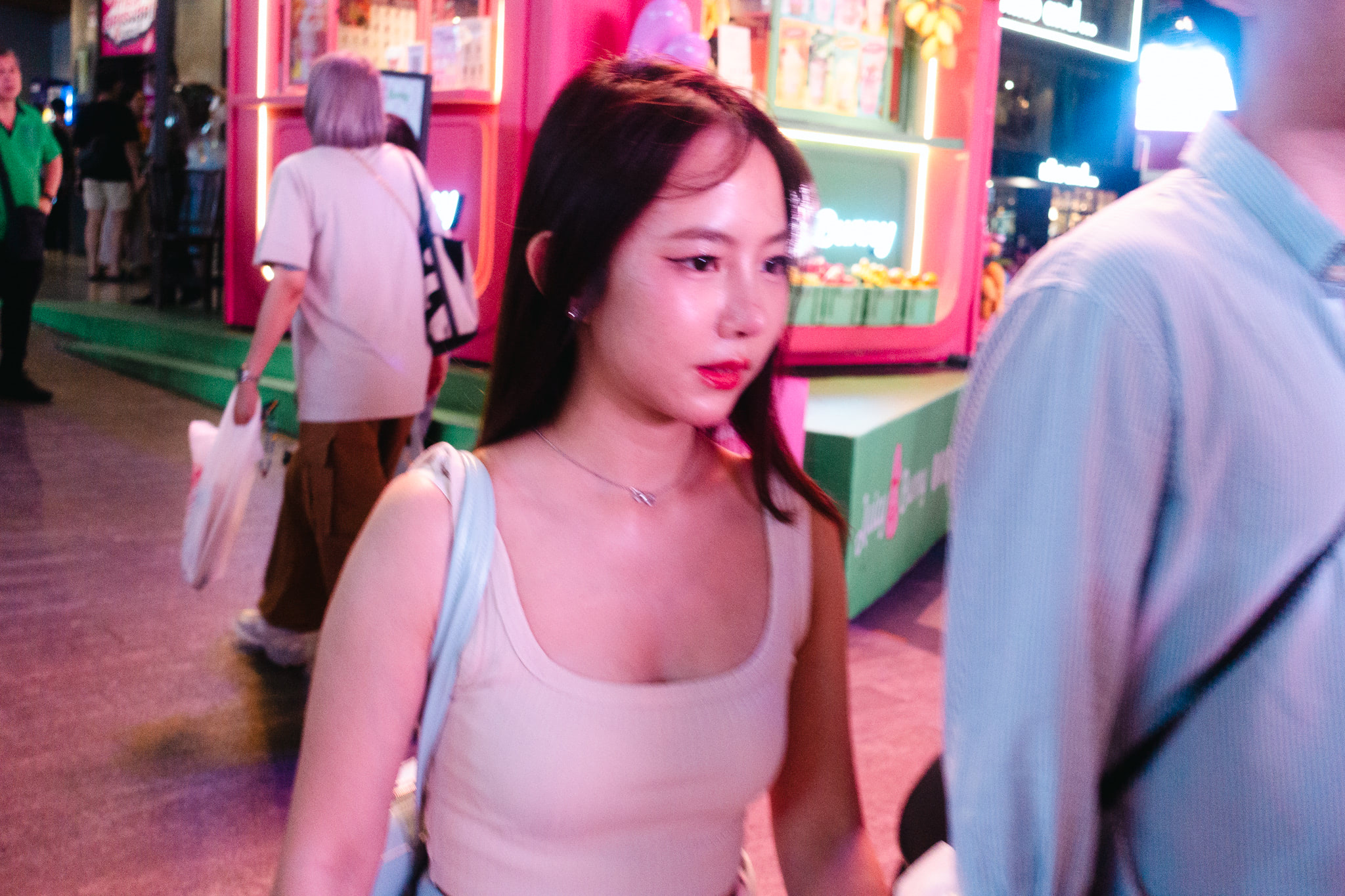 Young woman in a beige tank top walking past a brightly lit pink and green storefront.