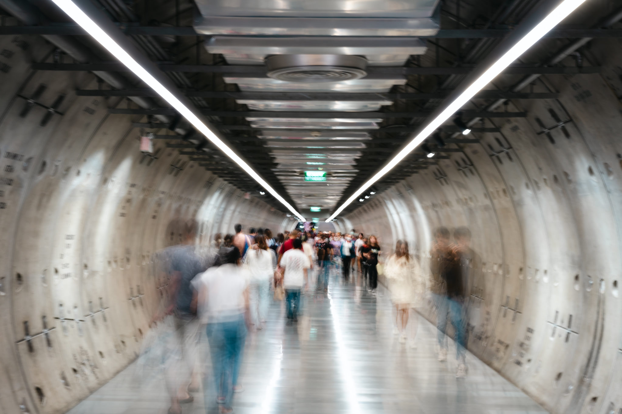 Blurred photo of many people walking through a long, brightly lit tunnel.