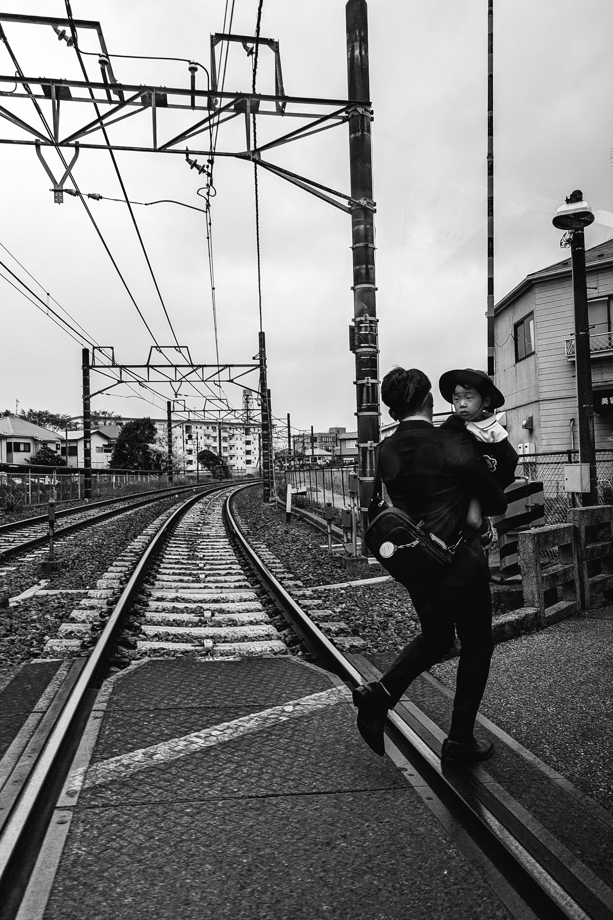 Black and white photo of a person carrying a child on their back, walking across railroad tracks.