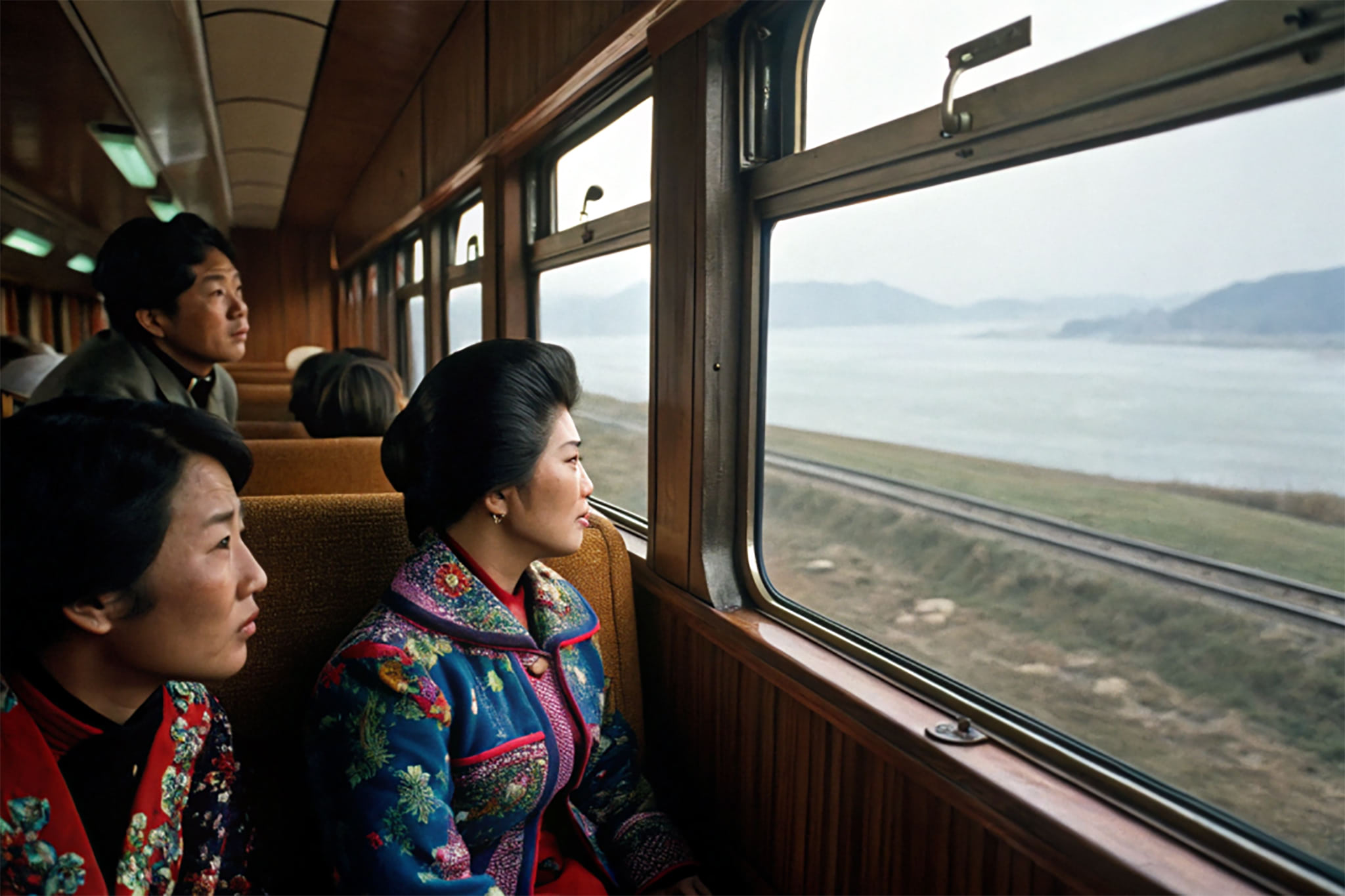 Three people sit on a train looking out the window at a body of water and mountains.