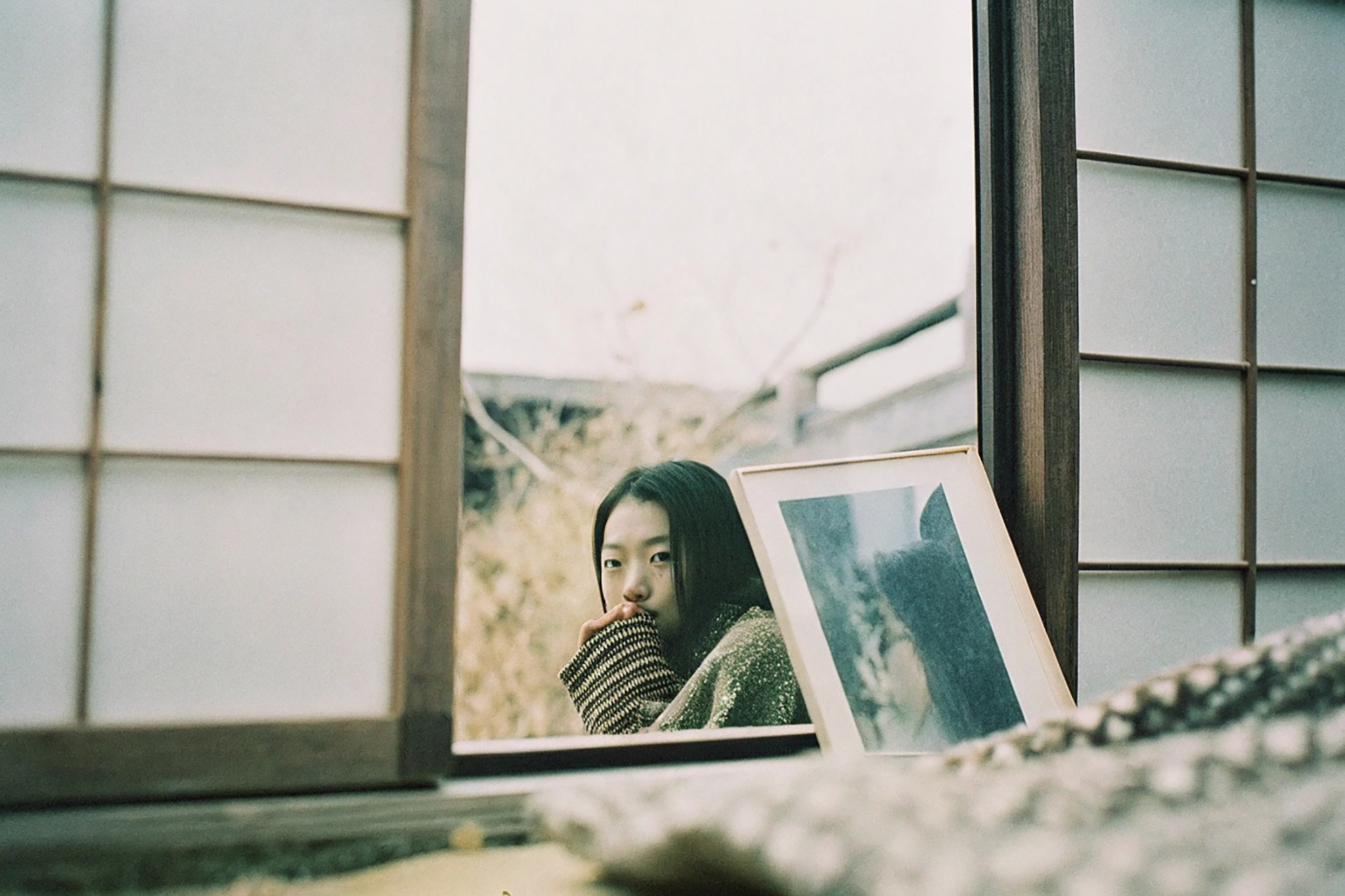 A young woman looks at the viewer from behind a window, hands covering her mouth.