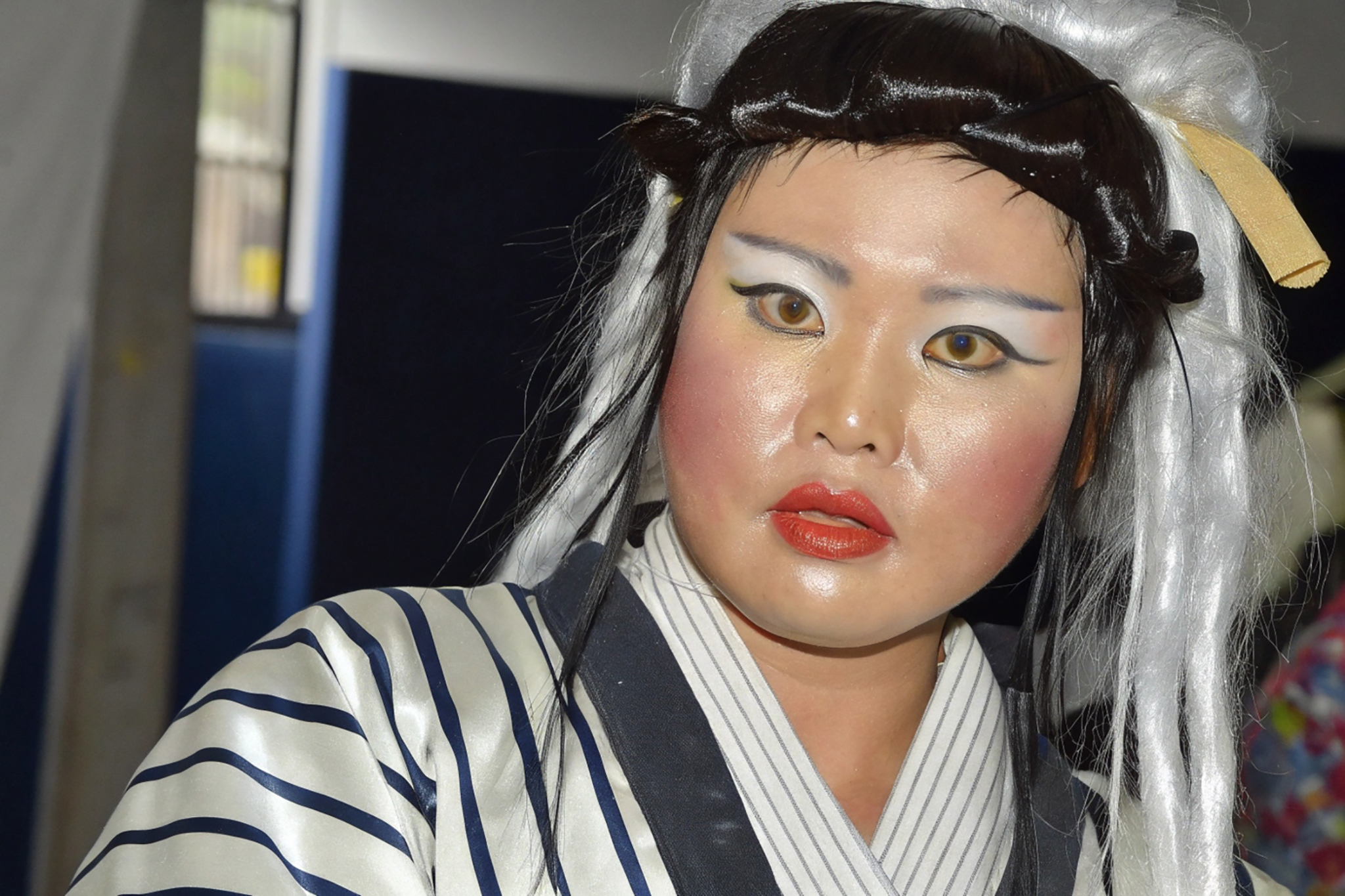 Close-up of a person in elaborate makeup and a striped kimono-style robe, wearing a styled wig with black and white hair.